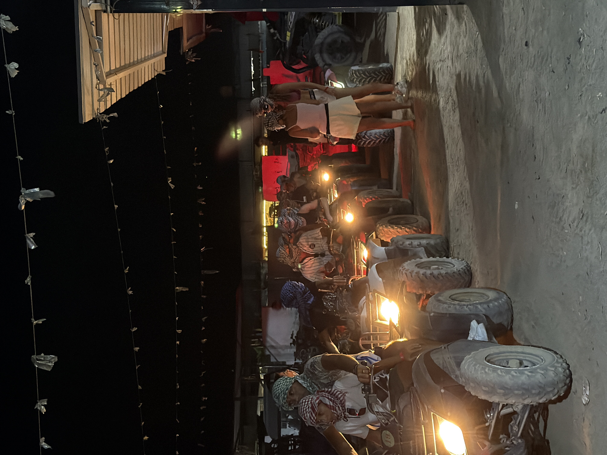 In Sharm El Sheikh, Egypt, at night, a group of people are lined up on ATVs in a dusty, outdoor area. The ATVs, mostly black and white, have their headlights illuminated, casting a bright glow. Many of the individuals are wearing traditional keffiyeh headscarves. The scene appears to be a staging area or starting point for an ATV tour. In the background, there is a simple building with a corrugated roof, decorated with strings of small, reflective objects. The overall atmosphere suggests a recreational activity taking place after dark.
