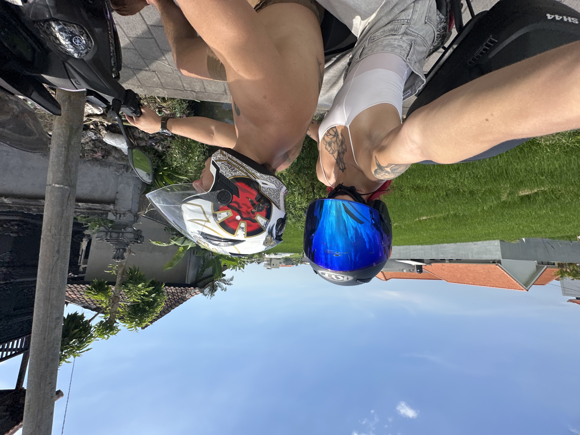 Two people wearing helmets sit astride a parked motorbike in North Kuta, Indonesia. The person on the left wears a white, red, and black helmet, and the person on the right wears a blue helmet. The background features buildings, a rice field, and a clear blue sky. A utility pole is visible on the left of the frame.
