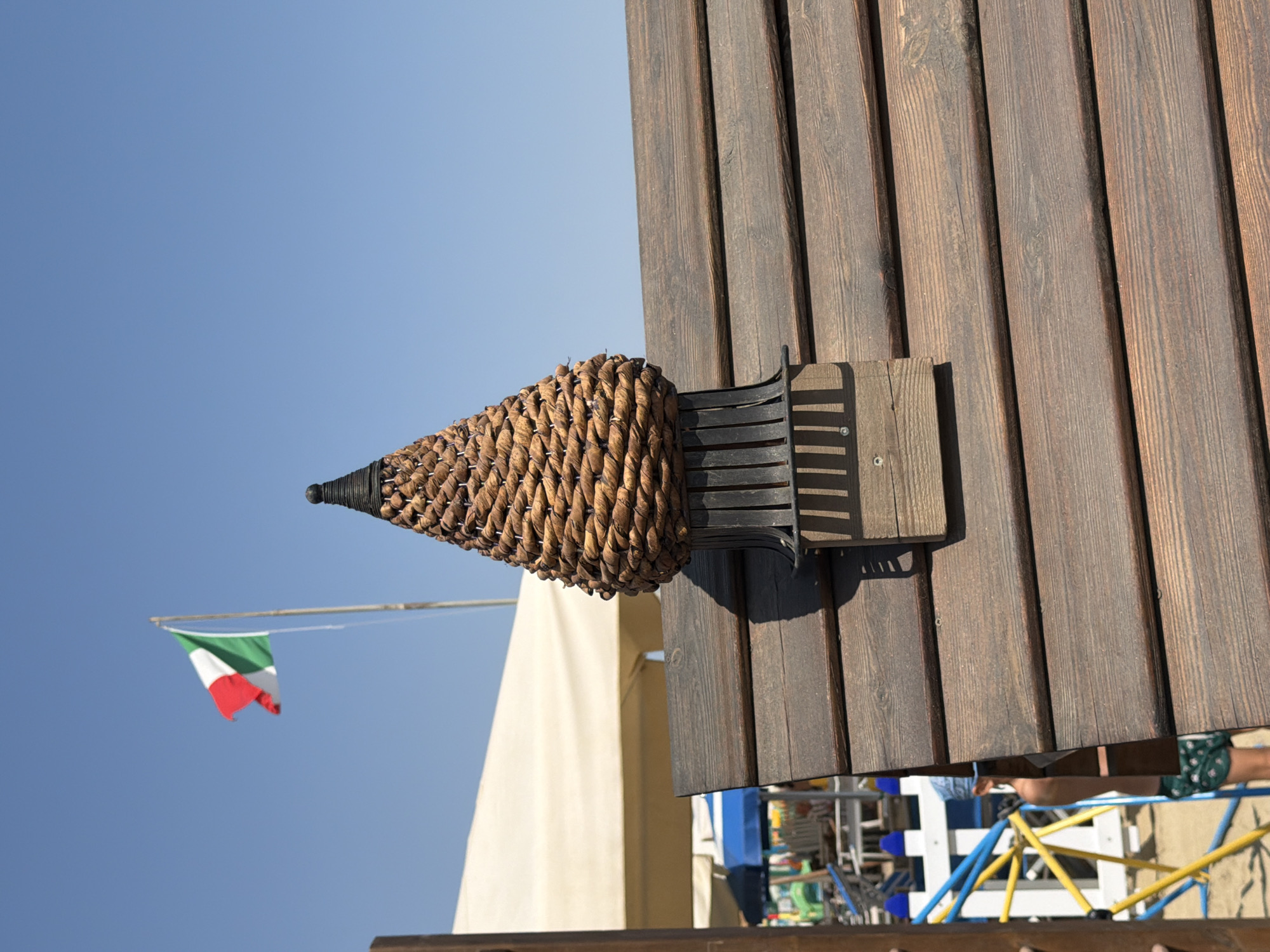 In Gatteo, Italy, a woven, cone-shaped ornament sits atop a dark wooden structure, likely part of a beach resort's decoration. The ornament features a dark cap and intricate weaving of natural fibers. Below, the weathered wooden planks of the structure are angled, creating a visual texture. In the background, the Italian flag flutters against a clear blue sky. Other beach elements are visible, including a large white canopy, beach umbrellas, and a colorful children's play structure. A person wearing a blue bandana and patterned shorts is partially visible, suggesting a recreational setting.