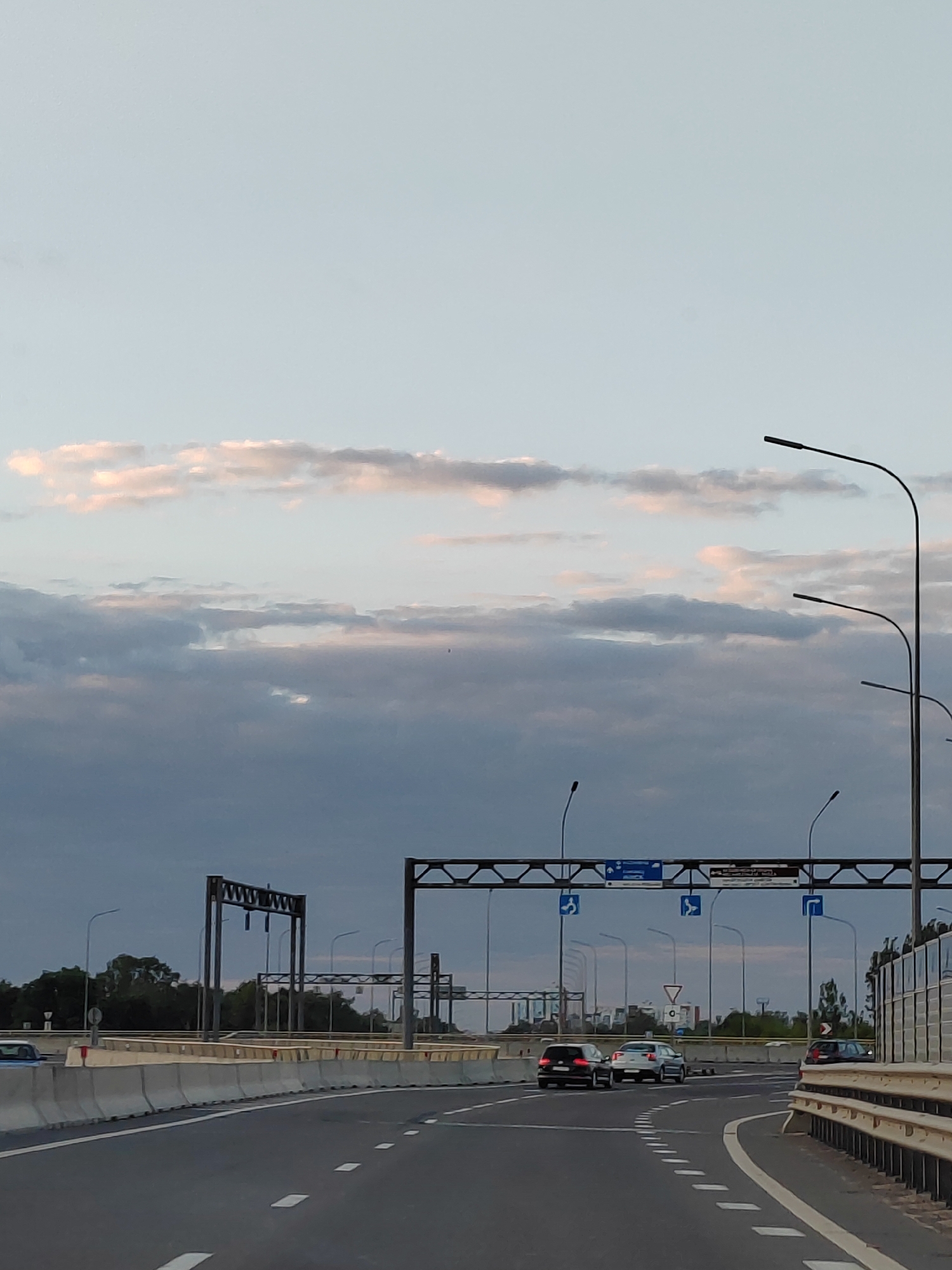 The image depicts a highway overpass in the city of Brest, Belarus, during twilight. The sky is overcast with a mix of gray and pale pink clouds. Several cars are visible on the road, including a black sedan, a silver hatchback, and a dark SUV. Overhead, there are road signs and streetlights, with structural supports for the overpass visible in the background. Concrete barriers line the side of the road, and a guardrail is present on the right side of the image. Trees and some buildings can be seen in the distance. The overall scene suggests a transportation hub within an urban environment during the evening hours.