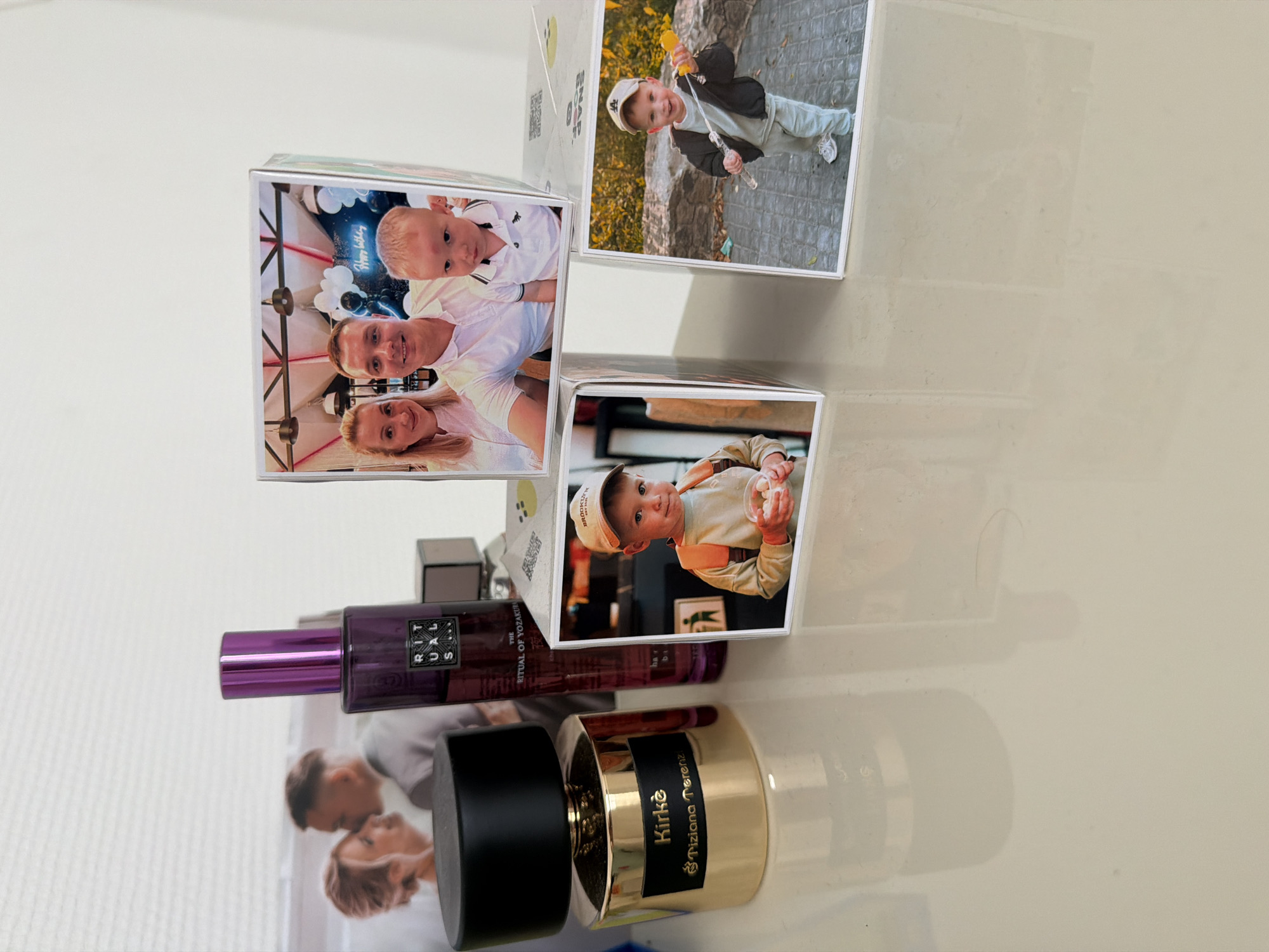A photograph taken from a slightly elevated angle on a flat, light-colored surface shows several objects, including perfume bottles and photo cubes. On the left, a gold perfume bottle with a black cap is visible. The label on the bottle reads "Kirkè" and "Tiziana Terenzi". Behind it, a tall, narrow purple bottle, possibly also perfume, stands upright with a metallic purple cap. The label on this bottle has text that is partially obscured but includes "RITUALS" and "YOZAKURA". To the right of the purple bottle, a small, dark grey rectangular object is present.
In the foreground, two photo cubes are arranged. The left cube displays a photograph of a young boy wearing a cap and an orange backpack, holding a white cup with small white objects inside. The right cube features a photograph of another young boy wearing a cap and a light-colored outfit, holding a toy that produces bubbles.
Behind the perfume bottles and photo cubes, partially visible in the background, is a large horizontal photograph or print. This image shows a couple, with the man leaning in towards the woman. The couple appears to be in a romantic pose, possibly at an event like a wedding.
The overall environment appears to be indoors, with a bright, white background, likely a wall. The surface on which the objects are placed is reflective, showing faint glints and shadows. There is no specific indication of the location being in Bila Tserkva, Ukraine, within the provided image.