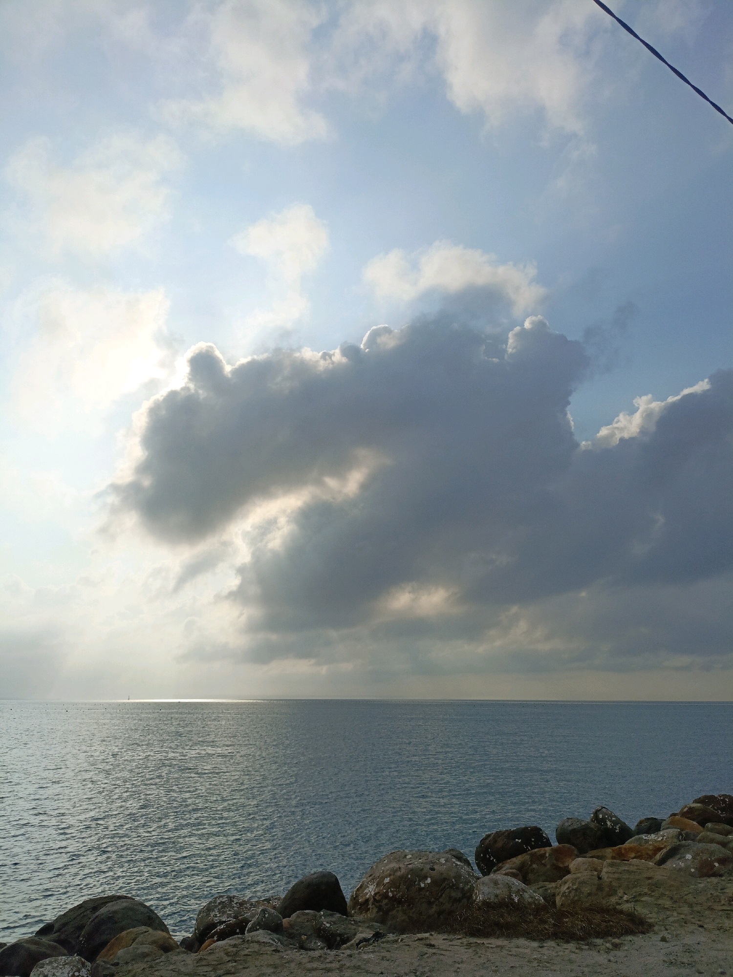 The image depicts a coastal scene with a wide expanse of blue ocean stretching towards the horizon. The water is slightly choppy, reflecting the light from the sky. A line of rocks and pebbles forms the foreground, leading the eye towards the sea. In the distance, a thin strip of land or a distant coastline is visible. The sky is a dramatic mix of blues and grays, with large, textured clouds dominating the upper portion of the frame. Sunlight breaks through a gap in the clouds, creating a bright, ethereal glow on the water's surface. A single dark line, possibly a wire, stretches diagonally across the upper right corner of the sky. The scene is likely located on the coast of Bulukumba, Indonesia, known for its stunning beaches and rocky shorelines.