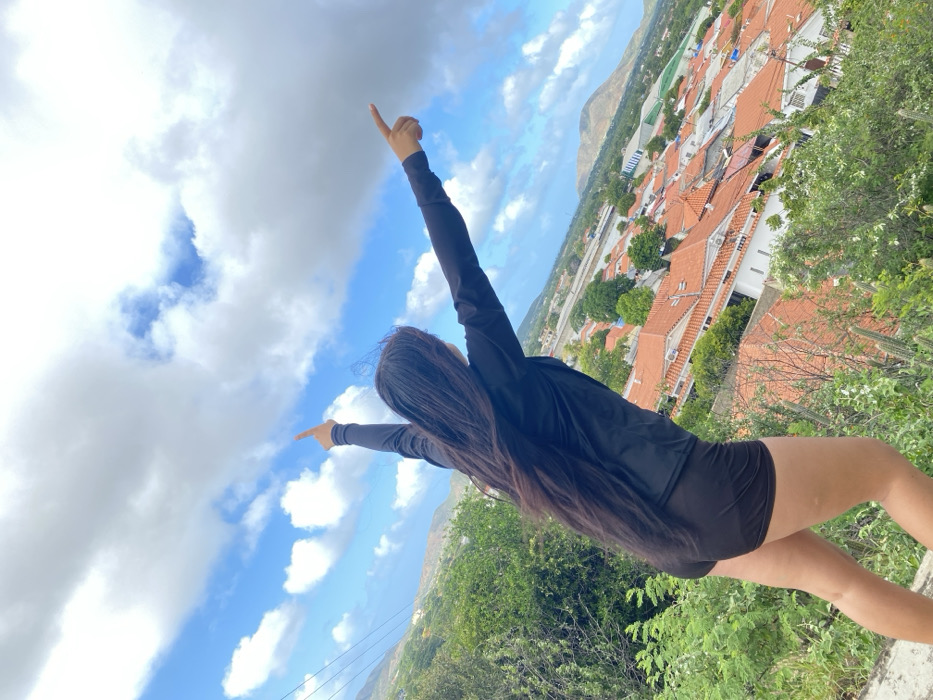 From a low angle, a woman stands with arms outstretched, pointing in opposite directions. She wears dark athletic clothing. Her long, dark hair hangs down her back. The background shows a landscape with red-roofed buildings nestled amongst trees and a distant mountain. The sky is filled with puffy white clouds against a blue expanse. Lush greenery frames the foreground.