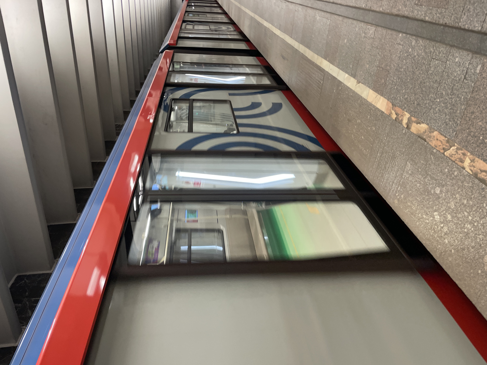 The image captures a Moscow Metro train at a station. The train is characterized by its red and blue exterior, and the white body is decorated with a distinctive blue concentric circle pattern. Reflections in the train's windows suggest the presence of passengers and station interiors. The scene includes a platform edge marked with a safety line and a wall with gray and brown stone tiles. Supporting columns are visible on the left side of the image. The blur indicates motion, suggesting that the train is moving or the photo was taken with a slow shutter speed.