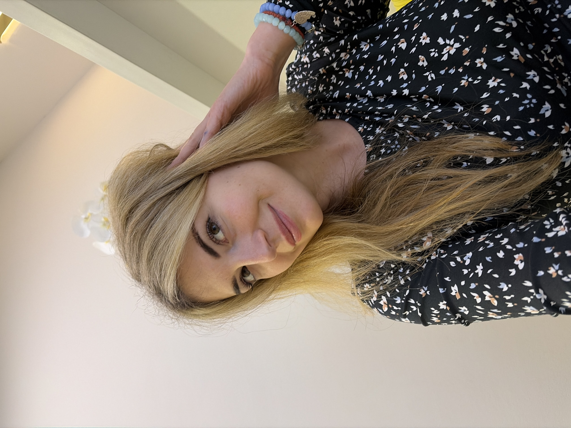 The image displays a head-and-shoulders portrait of a female individual in an indoor setting within Budva, Montenegro. The subject, appearing to be in her late 20s to early 30s, has long blonde hair and light skin, with greenish-brown eyes looking slightly off-camera to the viewer's left. A subtle smile is present on her face. She wears a black garment adorned with a repeating pattern of small white, light blue, and light brown floral shapes. Her right hand is raised, with fingers gently touching her hair near her right temple. On her right wrist, three beaded bracelets are visible: one light blue, one reddish-brown, and one dark blue, with a small metallic charm attached to the dark blue bracelet. The background consists of a plain, light-colored wall. In the upper right section, a light-toned architectural feature or shelf with integrated warm lighting is observed. A white orchid plant, displaying two blossoms, is partially visible in the upper background to the left of the architectural feature. The scene is evenly lit with warm artificial light.