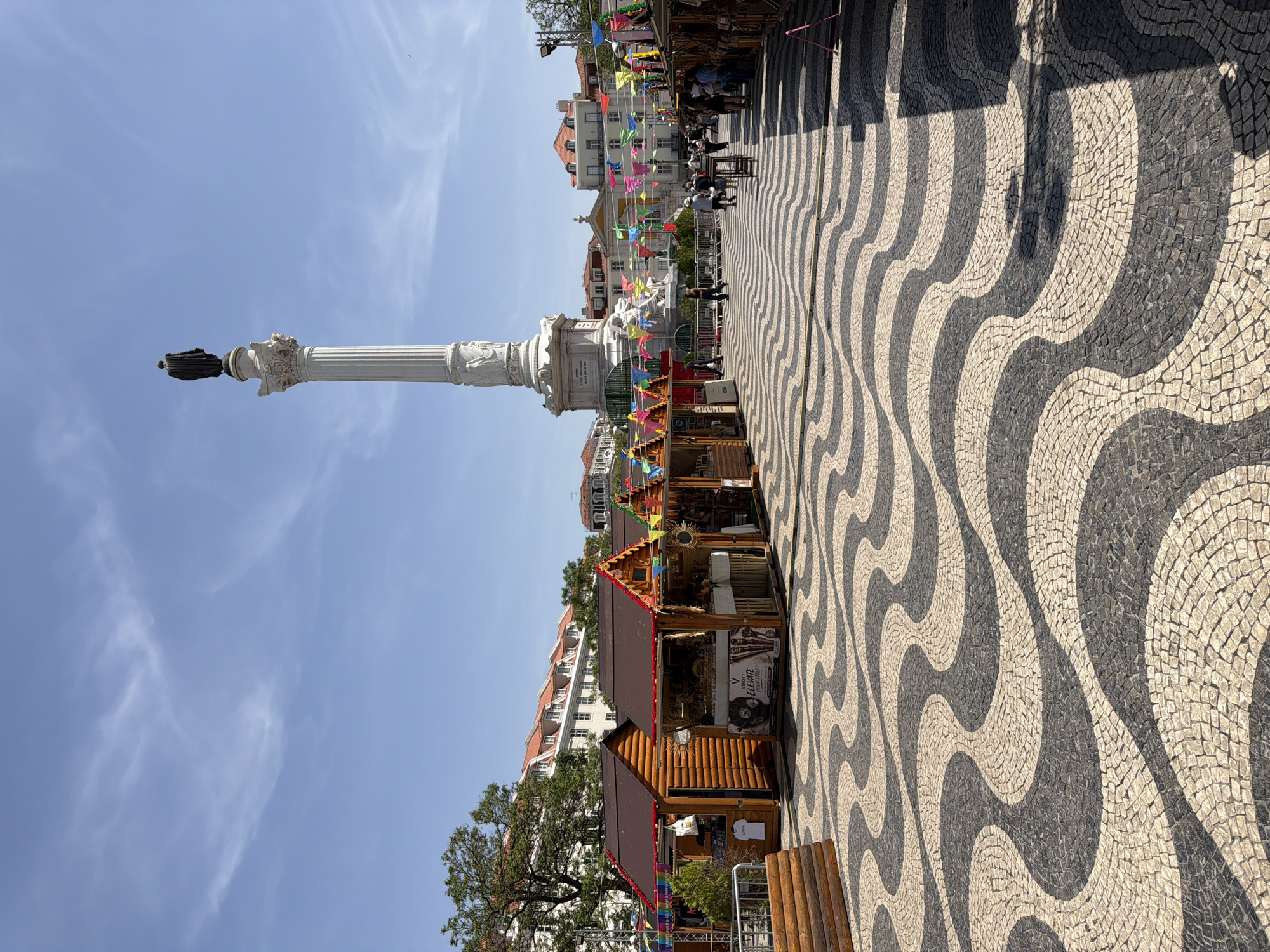 This image depicts a sunny day in Rossio Square (Praça do Rossio) in Lisbon, Portugal. The foreground is dominated by the iconic wave-patterned cobblestone pavement. Lining the sides of the square are temporary wooden market stalls decorated with colorful triangular flags. In the background, a tall white monument topped with a statue stands prominently against a clear blue sky with scattered white clouds. Several people are walking through the square, some appearing to be browsing the market stalls. Buildings with traditional tiled roofs are visible behind the monument and market stalls, characteristic of Lisbon's architecture. The overall atmosphere appears to be lively and festive.