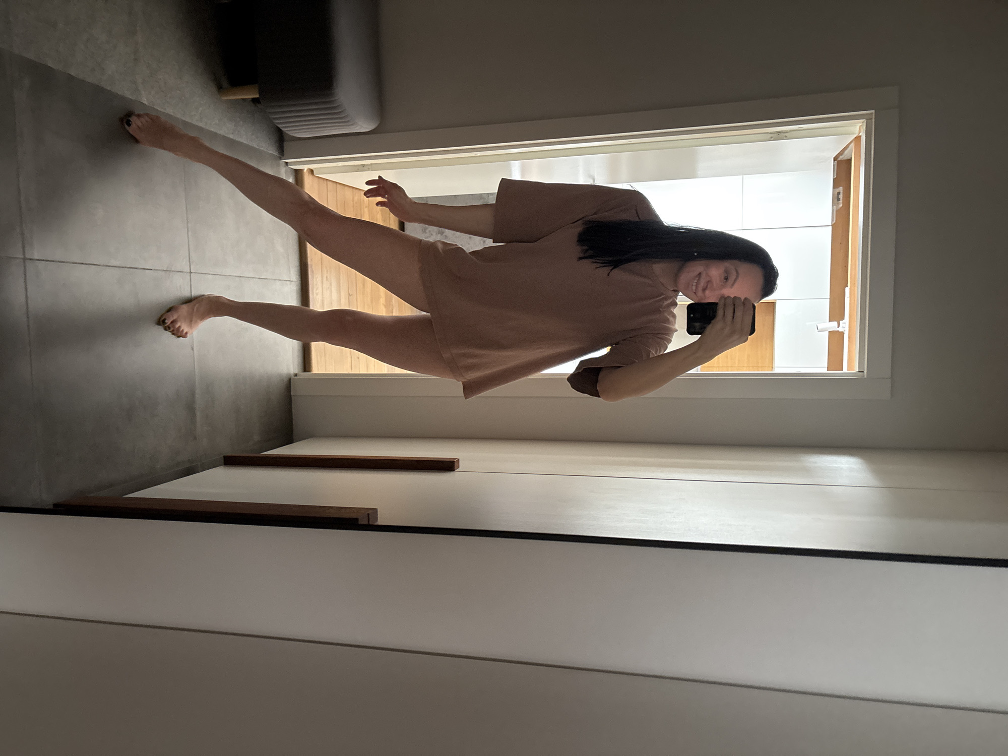 A woman is taking a selfie in what appears to be a bathroom. She is wearing a light brown t-shirt. Her dark hair is past her shoulders. She is barefoot, with her feet on the floor. The bathroom has gray tiled floors. In the background, there is a partially visible doorway leading to another room with white walls. A gray couch is partially visible in the upper left corner of the image. In the foreground, there is a white dresser with a dark trim.
I am unable to confirm the location as Chaikovsky, Russia, based on the image alone. There are no specific indicators of location.