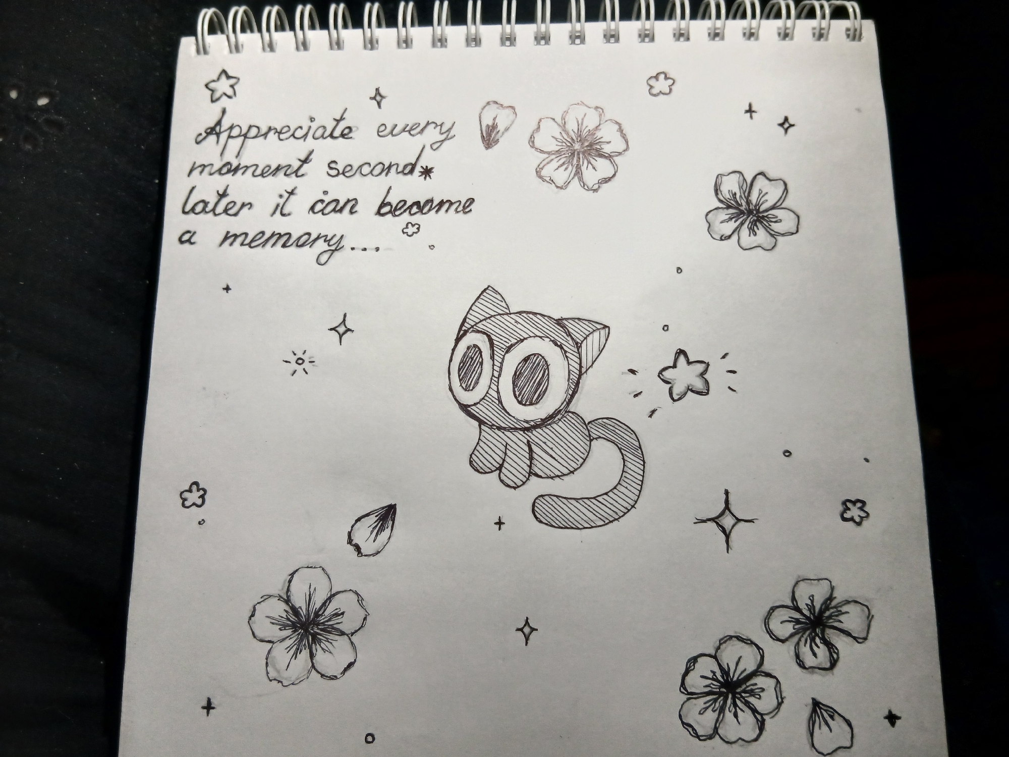 The image is a close-up of a page from a spiral-bound notebook. The page is filled with doodles drawn in black ink, including flowers, stars, and a stylized cat with large eyes. Handwritten text reads "Appreciate every moment second later it can become a memory..." The notebook rests on a dark surface. The location is not specified.