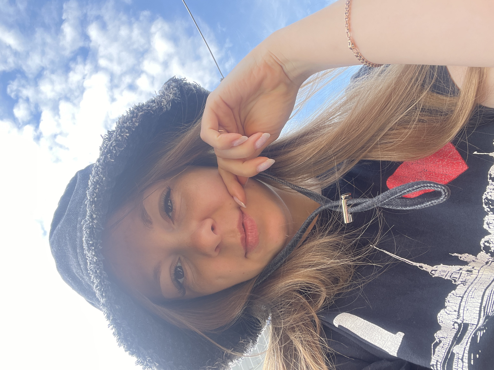 A young woman is photographed from a low angle, looking up at the camera. She is wearing a dark bucket hat with a frayed brim and a black hoodie with a graphic print of a city skyline and a red heart. Her hand is raised to her face, with her fingers touching her lips. She has long, blonde hair that falls around her shoulders. The background is a bright blue sky with scattered white clouds. The image appears to be taken outdoors on a sunny day. There are no specific details in the image that indicate the location is the city of Shadrinsk, Russia.