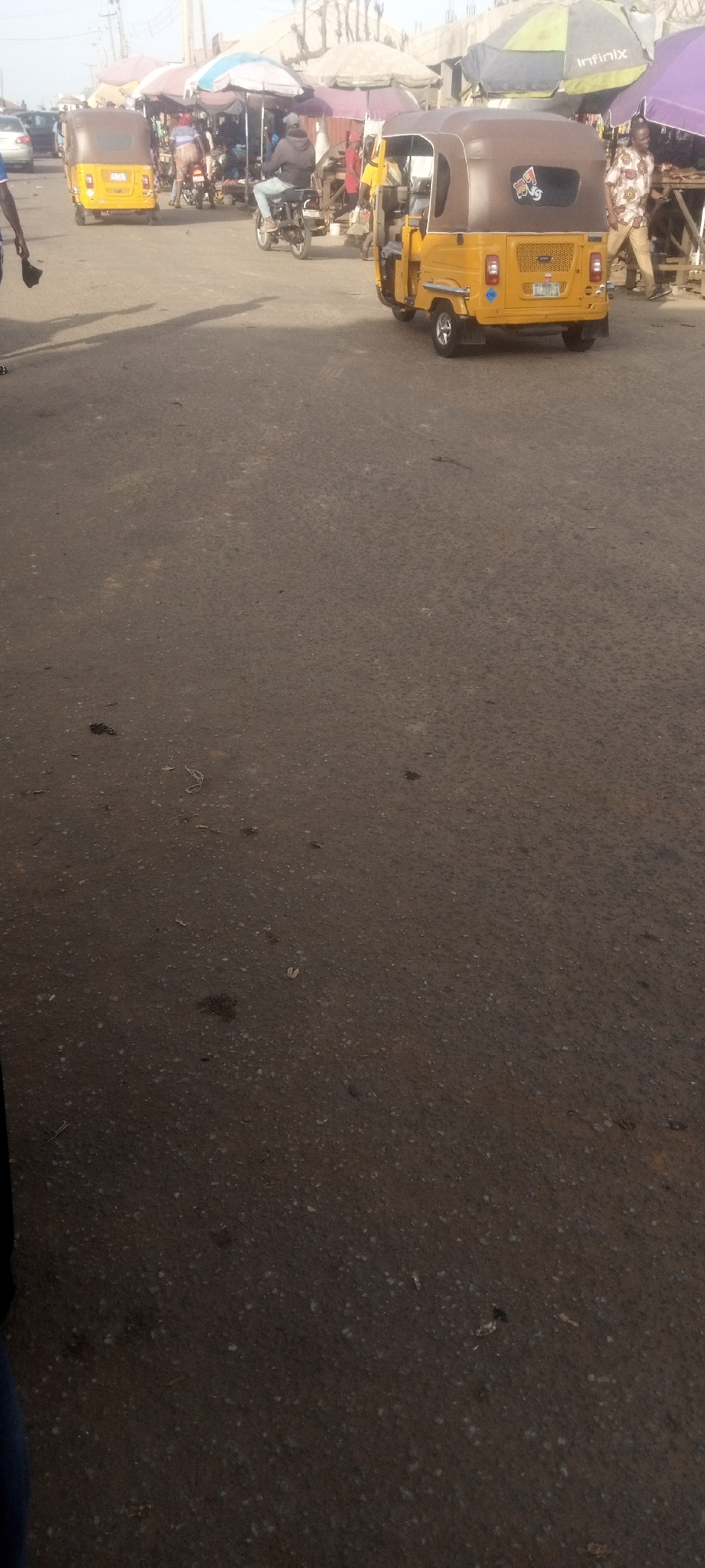 The image depicts a street scene in what appears to be the city of Narayi, Nigeria, populated with commercial tricycles (“Keke Napep”). There are pedestrians present and market umbrellas are visible. The scene seems to be centered around a marketplace or transit stop, with evidence of traffic and pedestrian activity. The road surface is dark and shows signs of wear.