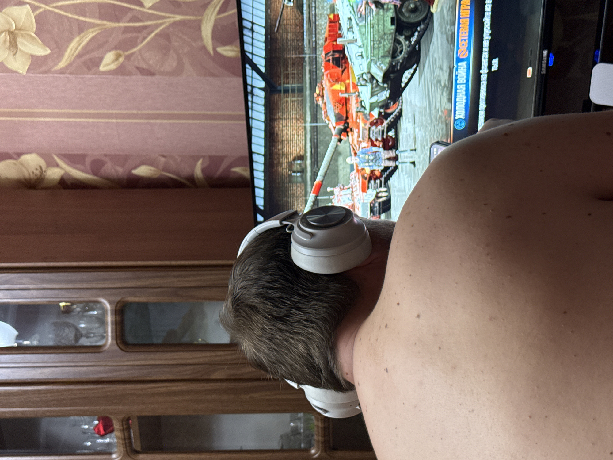 A shirtless person with short brown hair is facing a television screen while wearing white headphones. The television displays a video game featuring vehicles and text in a Cyrillic alphabet. Behind the person is a wooden cabinet with glass doors and a floral-patterned wallpaper. There is no evidence indicating the location of the image is Aksay, Kazakhstan.
