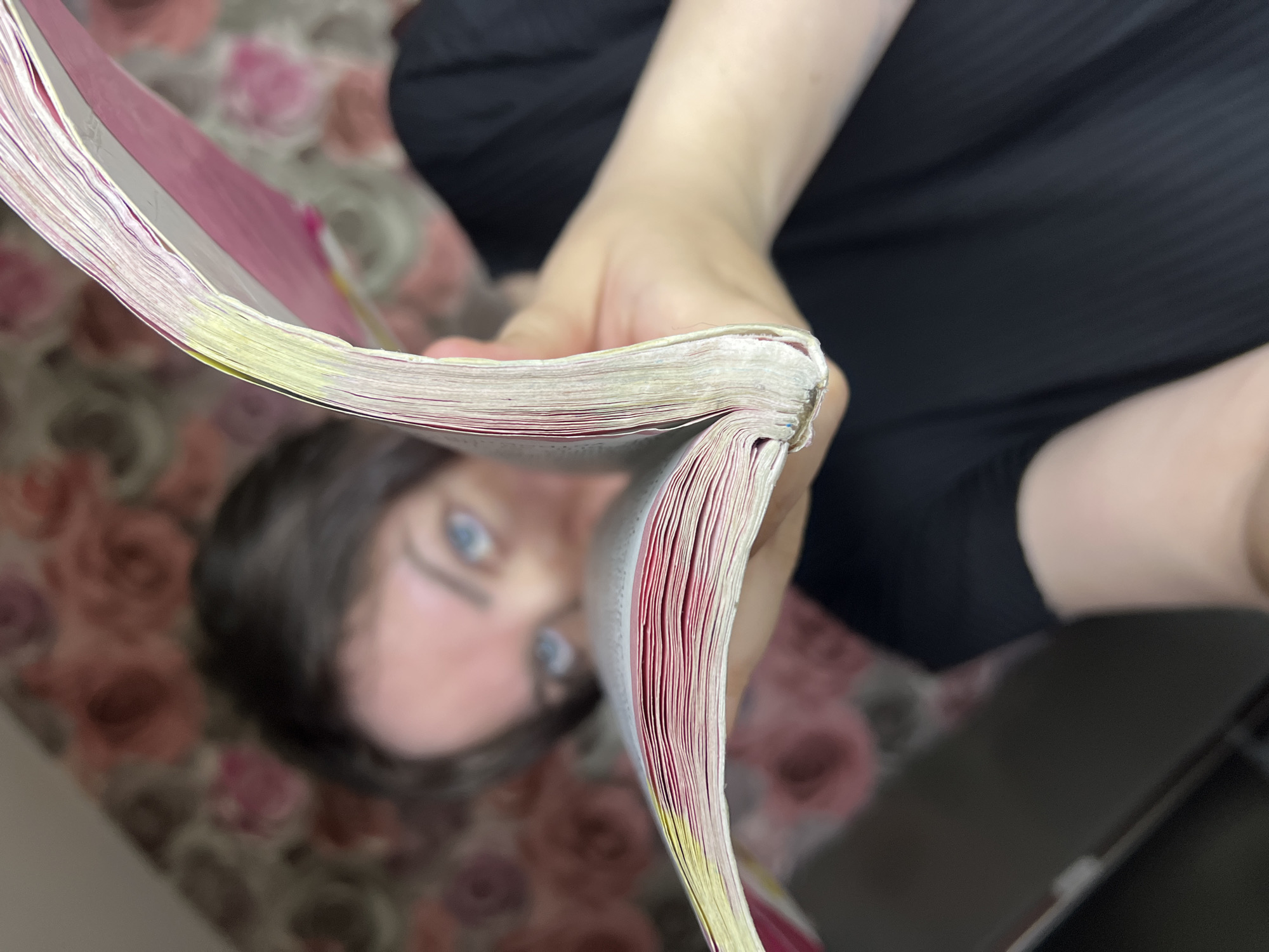 A person is holding open a thick book or magazine, presenting the pages to the viewer. The book appears well-used, with some pages marked with yellow highlighter. The person's face is visible behind the book, slightly out of focus, with blue eyes prominent. They are wearing a black shirt. The background consists of a floral-patterned fabric, possibly a bedspread or upholstery. No specific location is discernible, and there's no evidence to suggest the city of Noviy, Russia, is involved.