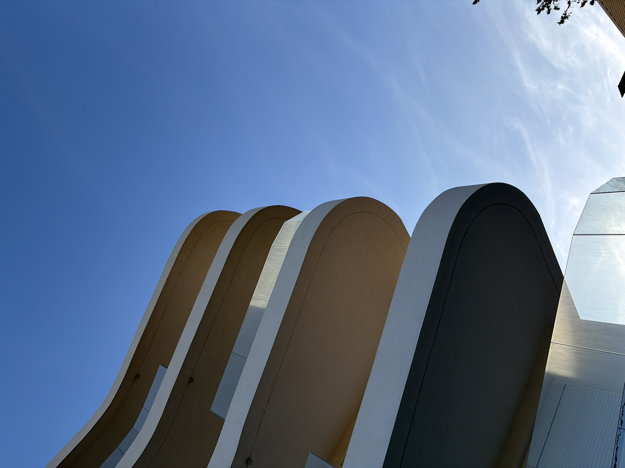 The image depicts an architectural structure with a series of curved, parallel fins or arches. The structures are white with tan-colored under surfaces and are set against a clear blue sky. The fins appear to be part of a building, and their repeating, rounded shapes create a sense of rhythm and movement. A section of the facade of the building with a combination of smooth and reflective material is seen in the right of the frame. A bit of foliage is present in the upper right corner. Considering the architectural style and the lack of information about the location, it is not possible to provide a more detailed description or confirm the location in the city of Municipal Unit of Elliniko, Greece.