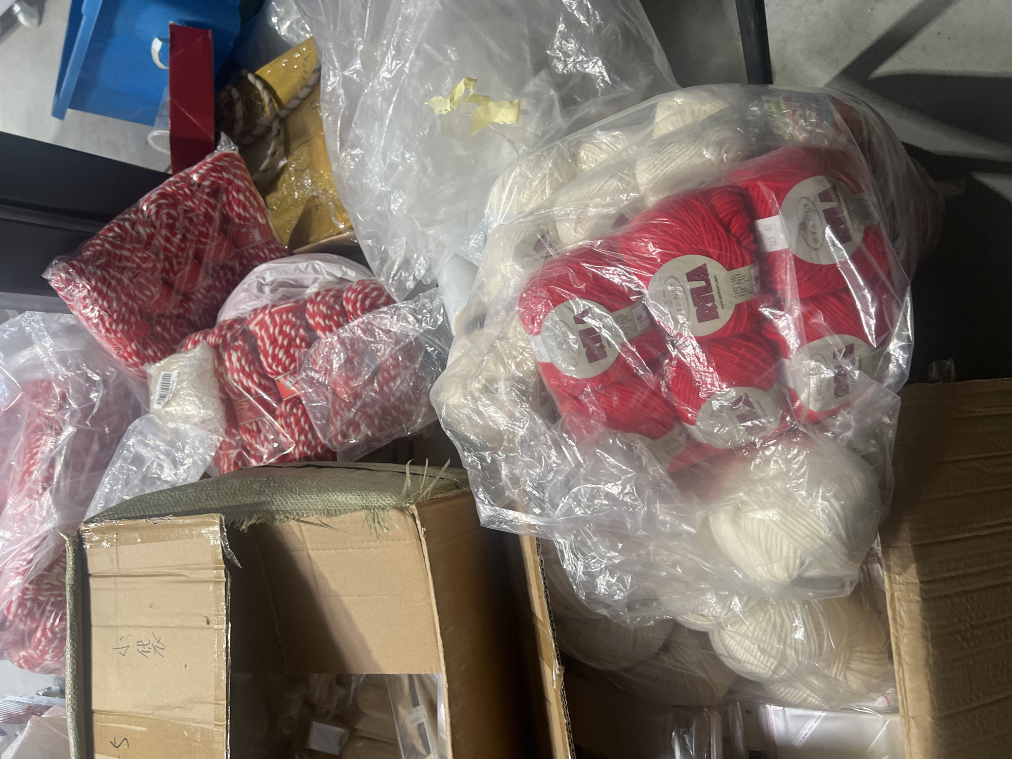 The image shows a cluttered storage area with boxes and bags filled with what appears to be yarn. The yarn is red and white, some in packages labeled "RILA." A cardboard box is partially open, revealing smaller items inside. The overall impression is of a storage or packing area, potentially in a warehouse or shop. There is no information to suggest this image was taken in Burgas, Bulgaria.