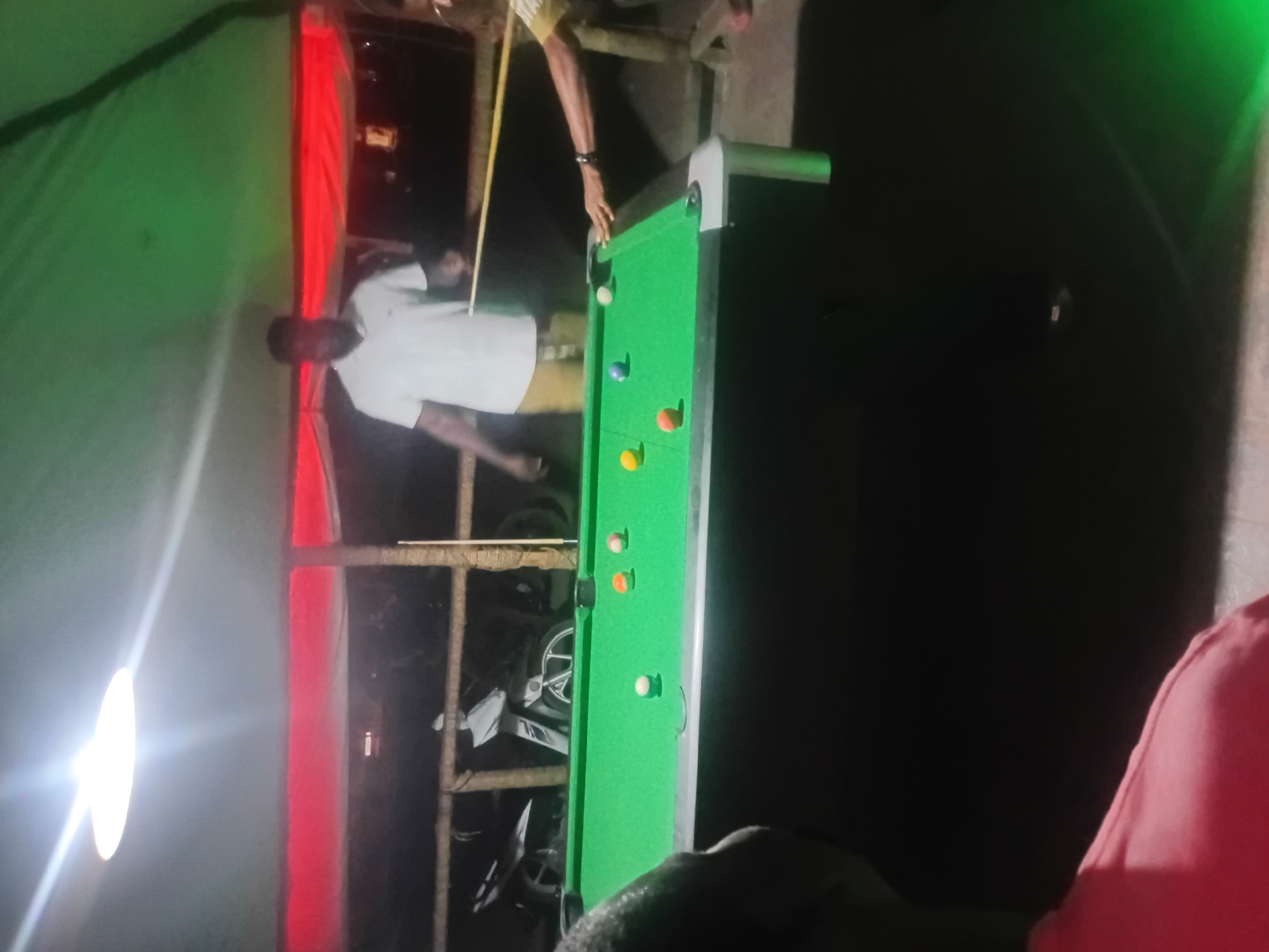 A man in a white shirt and khaki shorts is playing pool on a green pool table in Lokoja, Nigeria. There are several colored pool balls on the table. A cue stick is visible, presumably used by the man. In the background, there is a structure possibly made of wood, and a motorcycle is partially visible. The setting appears to be outdoors at night, under artificial lighting. A green and red structure can be seen to the left.