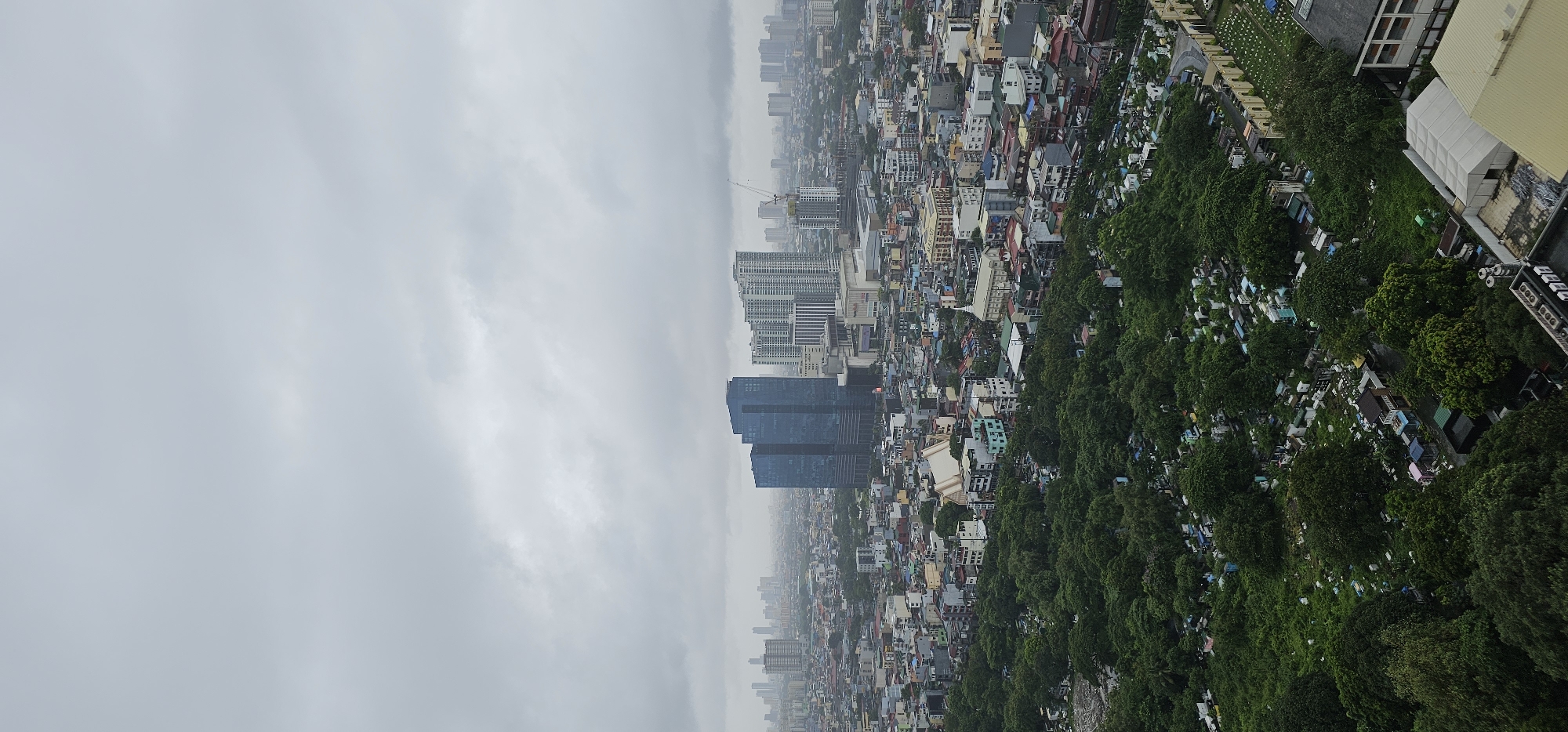 This is a high-angle, wide shot photograph of a dense urban cityscape under a heavily overcast sky. The image captures a mix of modern high-rise buildings and older, low-rise structures. In the foreground, a large expanse of lush green trees dominates the lower left quadrant, interspersed with what appears to be a cemetery or memorial park, evidenced by numerous small structures and pathways.
The midground reveals a sprawling residential and commercial area with buildings of varying heights and architectural styles, painted in a variety of colors. Two prominent, dark blue glass skyscrapers stand out in the center, with several lighter-colored, multi-story residential buildings to their right. A construction crane is visible near the residential towers, indicating ongoing development. In the distance, a hazy outline of more skyscrapers and urban sprawl stretches towards the horizon.
The overall atmosphere is muted due to the cloudy weather. The location is identified as Makati, Philippines. There are no discernible people or specific actions depicted in the scene; the focus is on the urban landscape and its components. The composition emphasizes the scale and density of the city, contrasted with the natural element of the green space in the foreground.