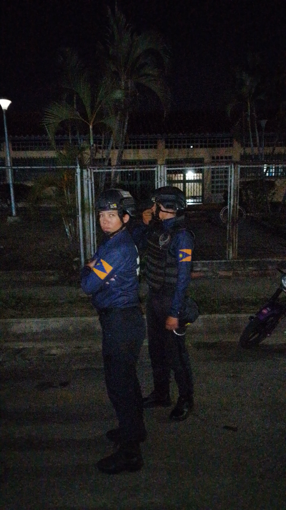 Two individuals wearing dark clothing, protective helmets, and tactical vests are standing on a dark paved surface at night. Both individuals have a blue sleeve with a yellow chevron. One of the individuals has their arms crossed. In the background, there is a building with a fence, a lamppost, and trees. The location is Tinaquillo, Venezuela.