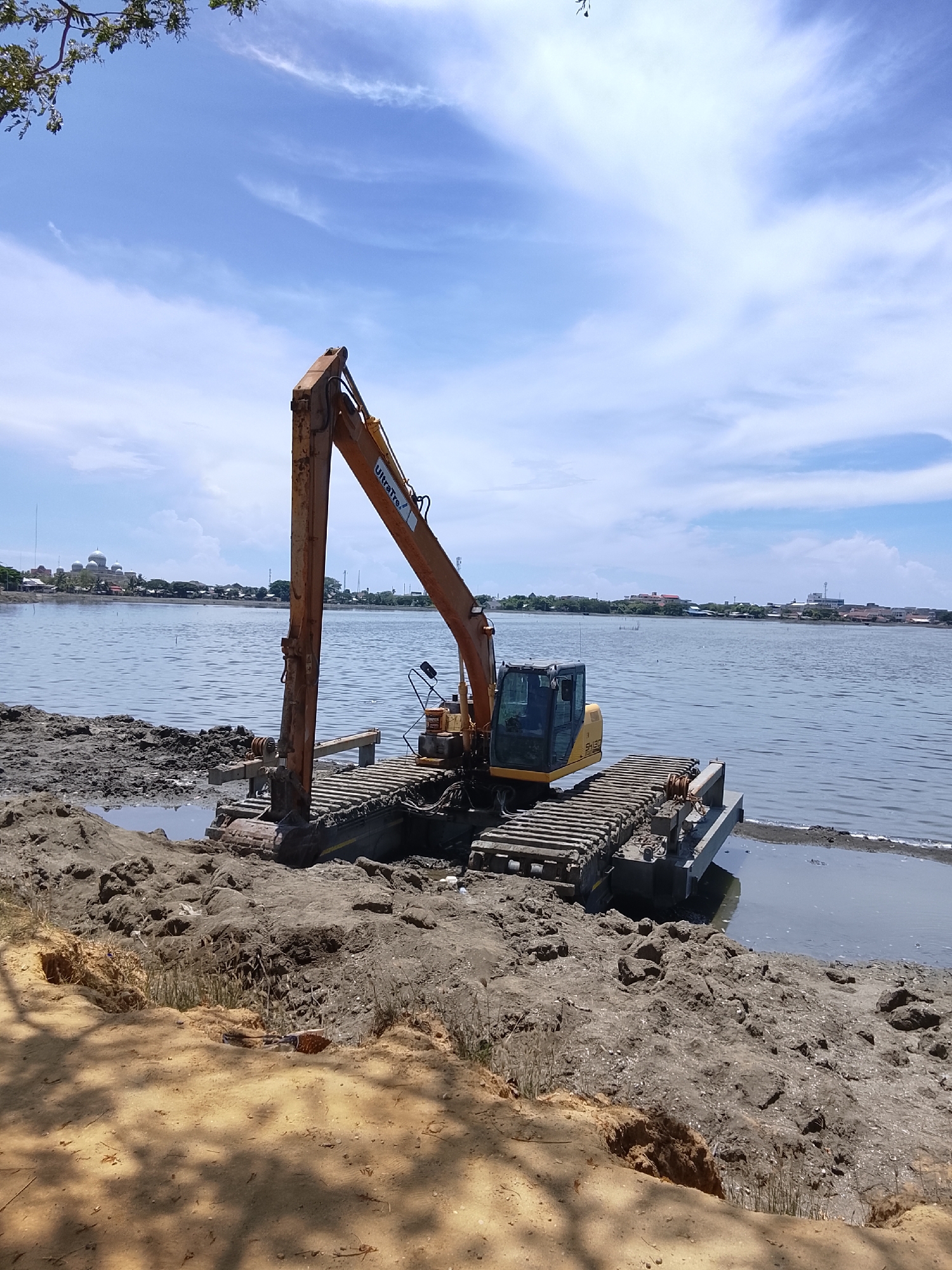 An amphibious excavator with a long orange boom is parked on a muddy shoreline, with its tracks partially submerged in the water. The excavator's cabin is yellow, and the machine appears to be engaged in dredging or land reclamation work. The foreground consists of disturbed soil and sand with some vegetation. In the background, a wide body of water stretches to the horizon, where a city skyline is visible. The city features buildings, including a prominent mosque with a large dome, characteristic of Lhokseumawe, Indonesia. The sky is blue with scattered white clouds. The image captures a construction or infrastructure project taking place near the coast in a populated area.