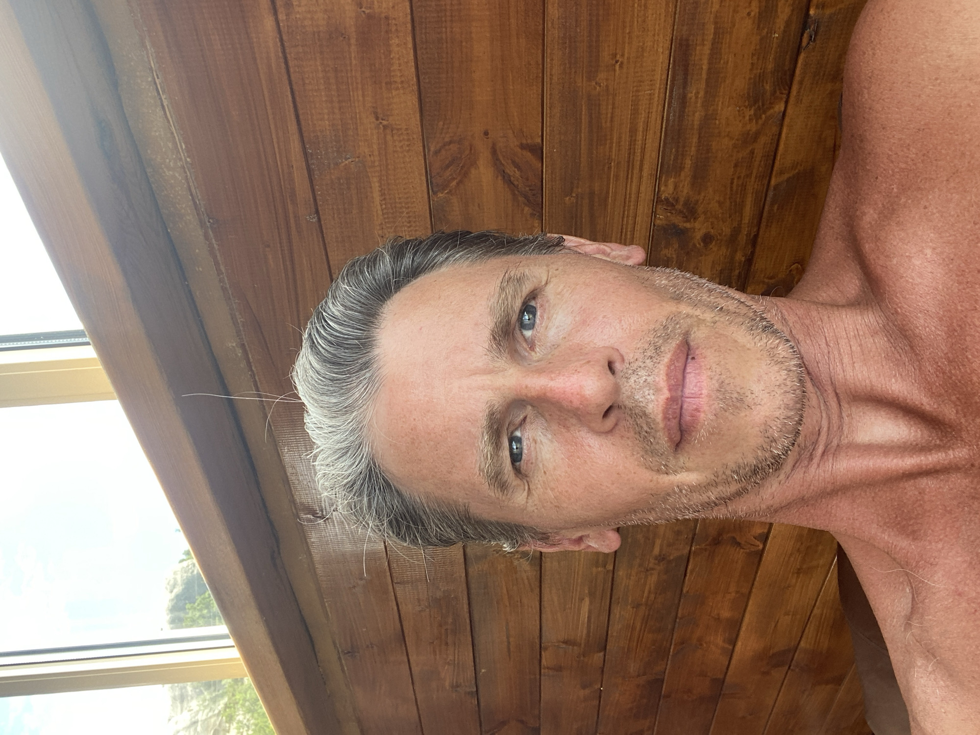 A close-up, slightly upward angled selfie shows a man's head and shoulders. He has graying hair slicked back, blue eyes, and a light stubble beard. His skin appears sun-kissed and slightly red, with visible texture and wrinkles around his eyes and neck. The background consists of horizontal wooden planks, with a window and a glimpse of sky and rocks visible at the top. The scene appears to be indoors, possibly in a rustic cabin or sauna, given the wooden interior and natural light. No specific location within Krasnomakskoye Rural Settlement, Ukraine can be determined from the visual evidence.