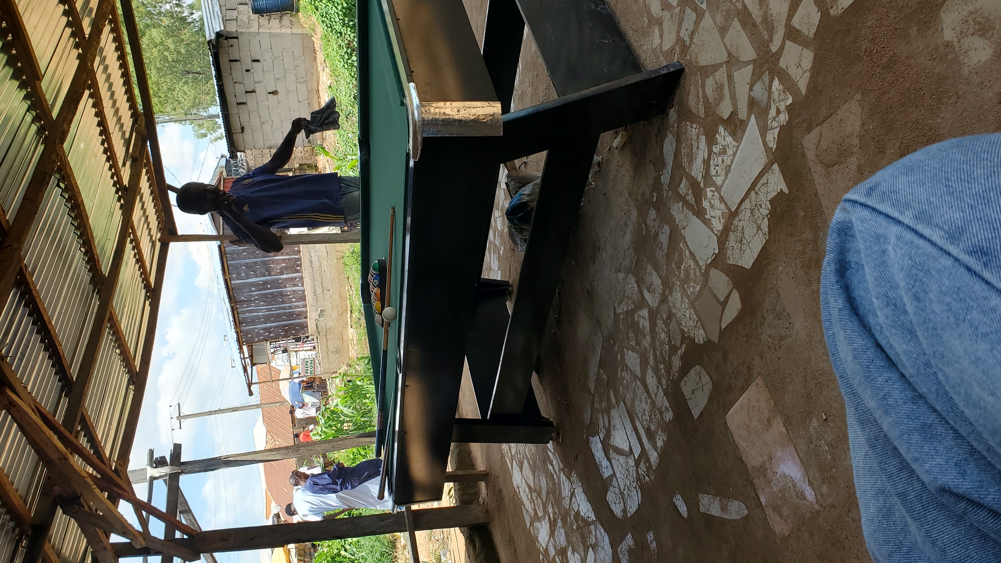 The image depicts an outdoor pool table setup, possibly in a local gathering spot within Jalingo, Nigeria. A man in a blue t-shirt and jeans stands near the table, holding a black cloth in his right hand. He appears to be interacting with the pool table area. Several billiard balls, including a white cue ball and colored balls, are visible on the green felt surface of the table, along with a pool cue. The table itself is dark colored and supported by a sturdy frame. The ground beneath is a mixture of soil and scattered, irregular white tiles, suggesting a somewhat rustic setting. In the background, other individuals are partially visible, some wearing white attire, and a few simple buildings with corrugated metal roofs can be seen. Lush green vegetation, including what appears to be corn stalks, lines the background, indicating an area that might be on the outskirts of a more built-up part of the city. The scene is covered by a roof supported by wooden beams, providing shade from the sun. The overall impression is of a casual, communal space for recreation.