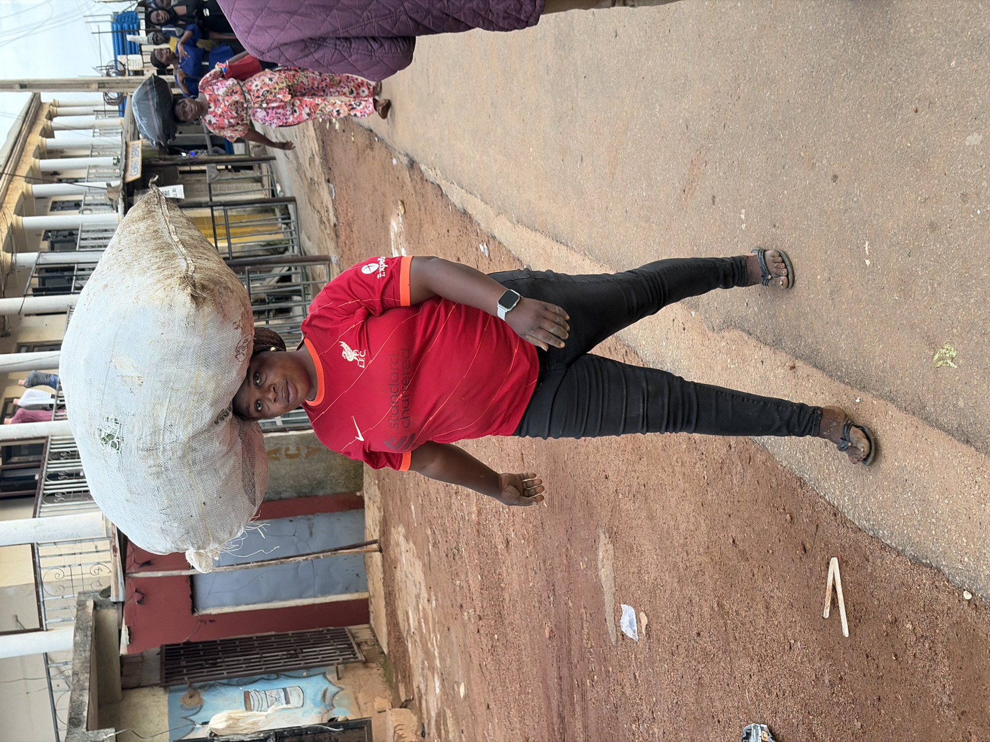 A woman in a red Liverpool F.C. jersey and black pants walks on a dirt and paved road in Gura Top, Nigeria, carrying a large white sack on her head. She is wearing sandals. The road is lined with buildings, some with balconies and columns. Other people are visible in the background, some also carrying items on their heads. The ground is a mix of reddish-brown dirt and grey pavement. The sky is overcast.
