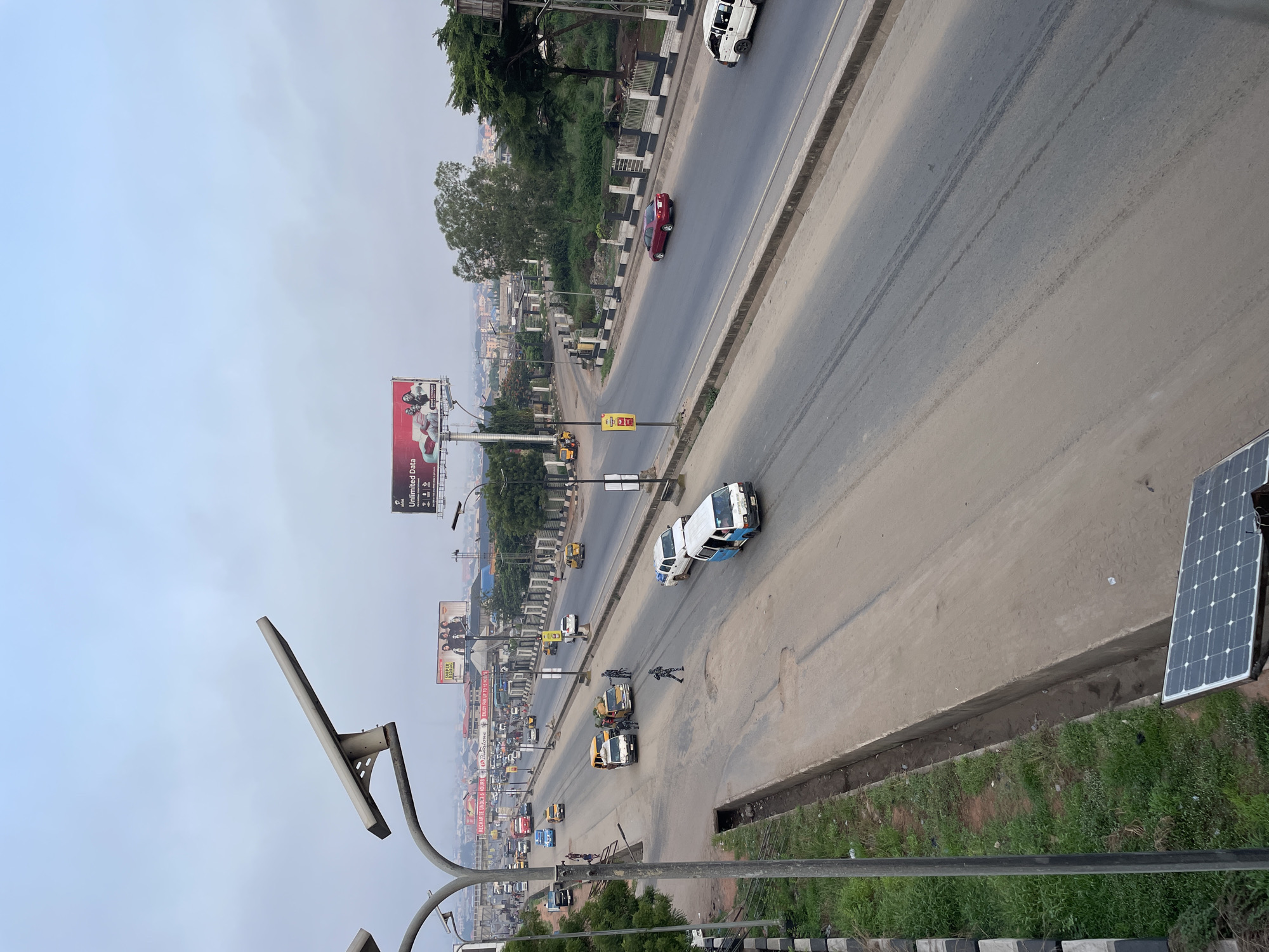 The image shows an elevated view of a bustling street in Onitsha, Nigeria. Traffic is visible on the road, including cars, vans, and small trucks. Several pedestrians are walking alongside the road. Numerous billboards are erected along the road. The sky is overcast, casting a diffused light. The foreground includes a lamppost and a solar panel, suggesting a focus on renewable energy. Vegetation grows along the roadside.