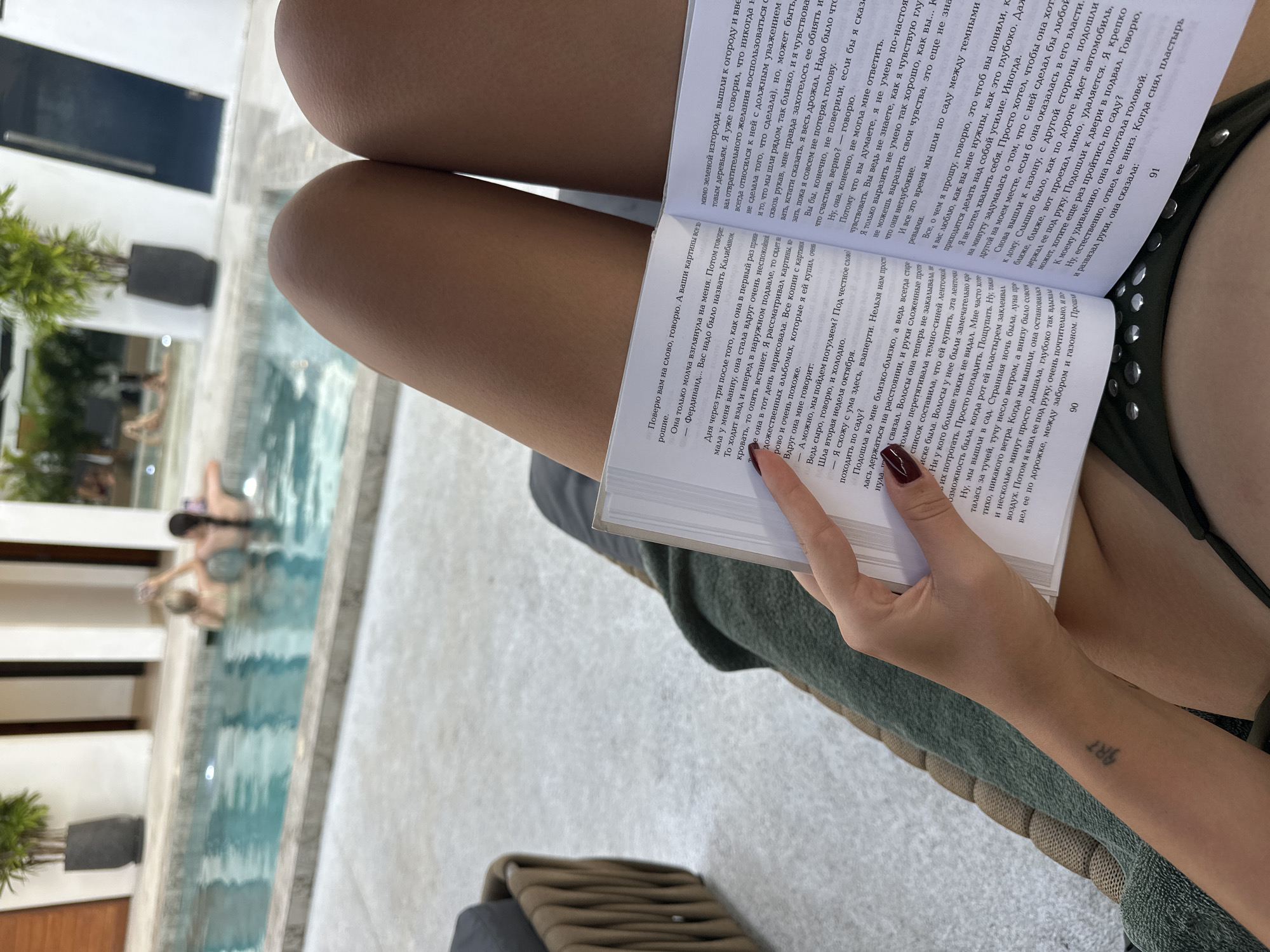 A first-person perspective shot shows a person's legs and hands holding an open book. The book is in Russian. The person is sitting on a lounge chair covered with a green blanket. In the background, out of focus, is a swimming pool with two people in it. The environment appears to be an outdoor patio with modern architecture. The location is the city of Si Sunthon, Thailand.