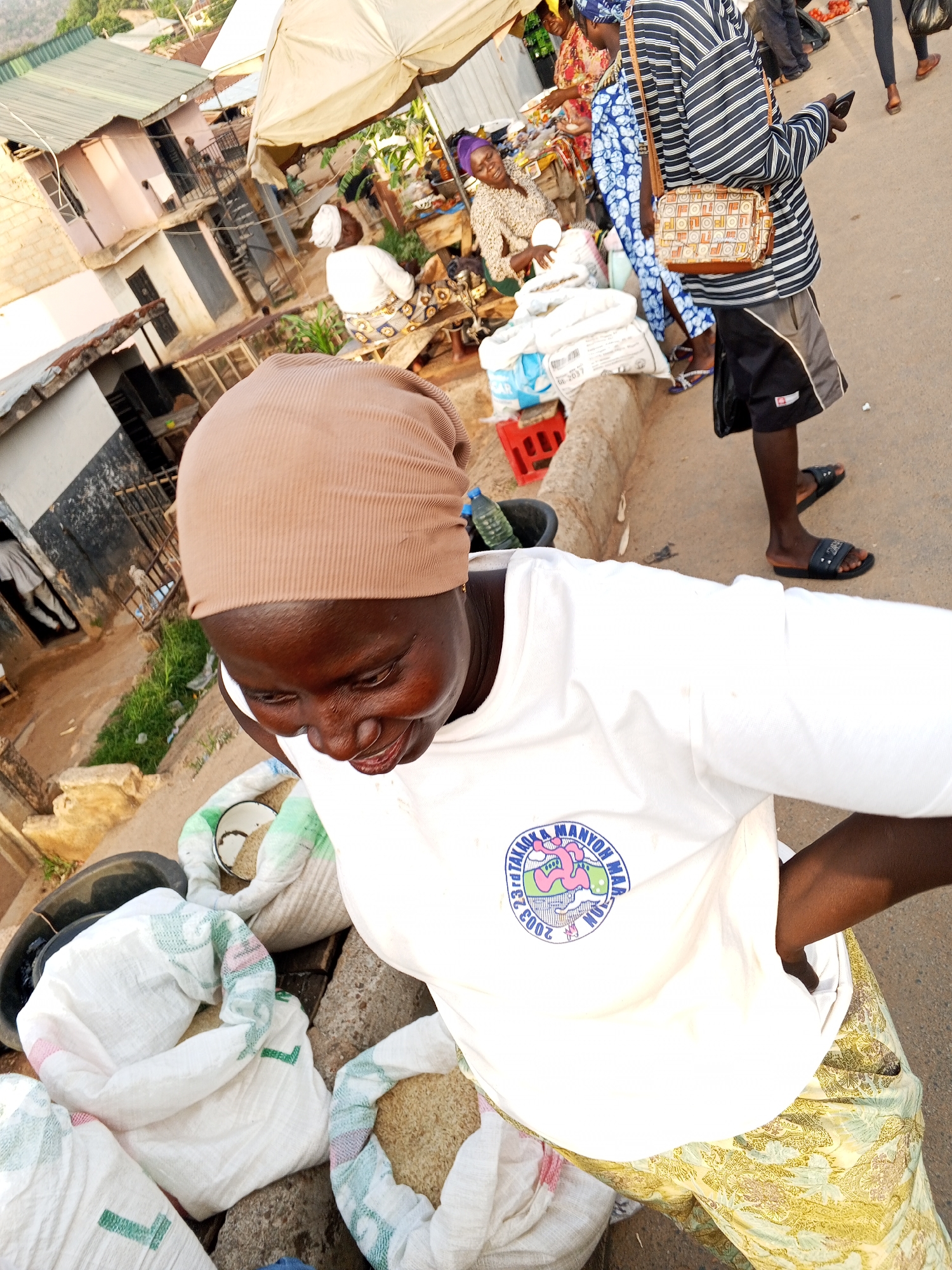 The image shows a street market scene in Gwong, Nigeria. A woman wearing a tan head covering and a white t-shirt with a logo is shown close to the camera, with bags of grain or rice near her. Behind her, there are other vendors and customers, some under an umbrella. One man in a striped shirt is walking past. Buildings are visible in the background. The setting appears to be a bustling area with market activity taking place.