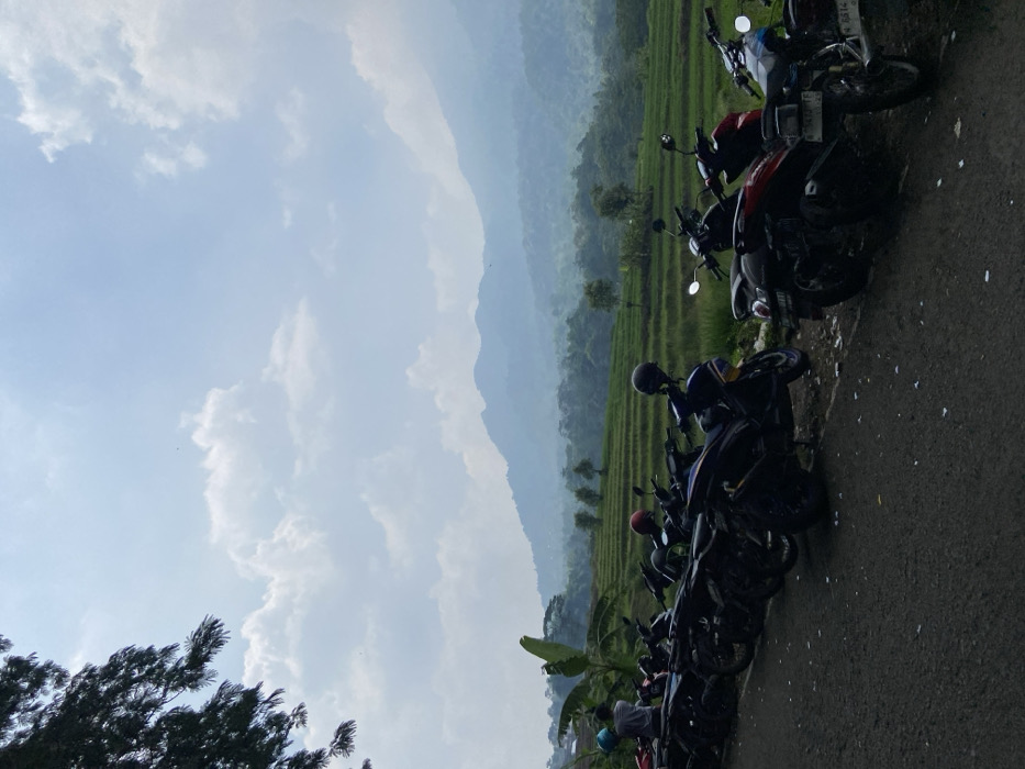A landscape photograph shows a row of motorcycles parked alongside a road with lush green rice terraces in the background. The sky is partly cloudy with visible mountains in the distance obscured by a light haze. Trees and shrubs are present near the road. The image suggests a rural or semi-rural setting.