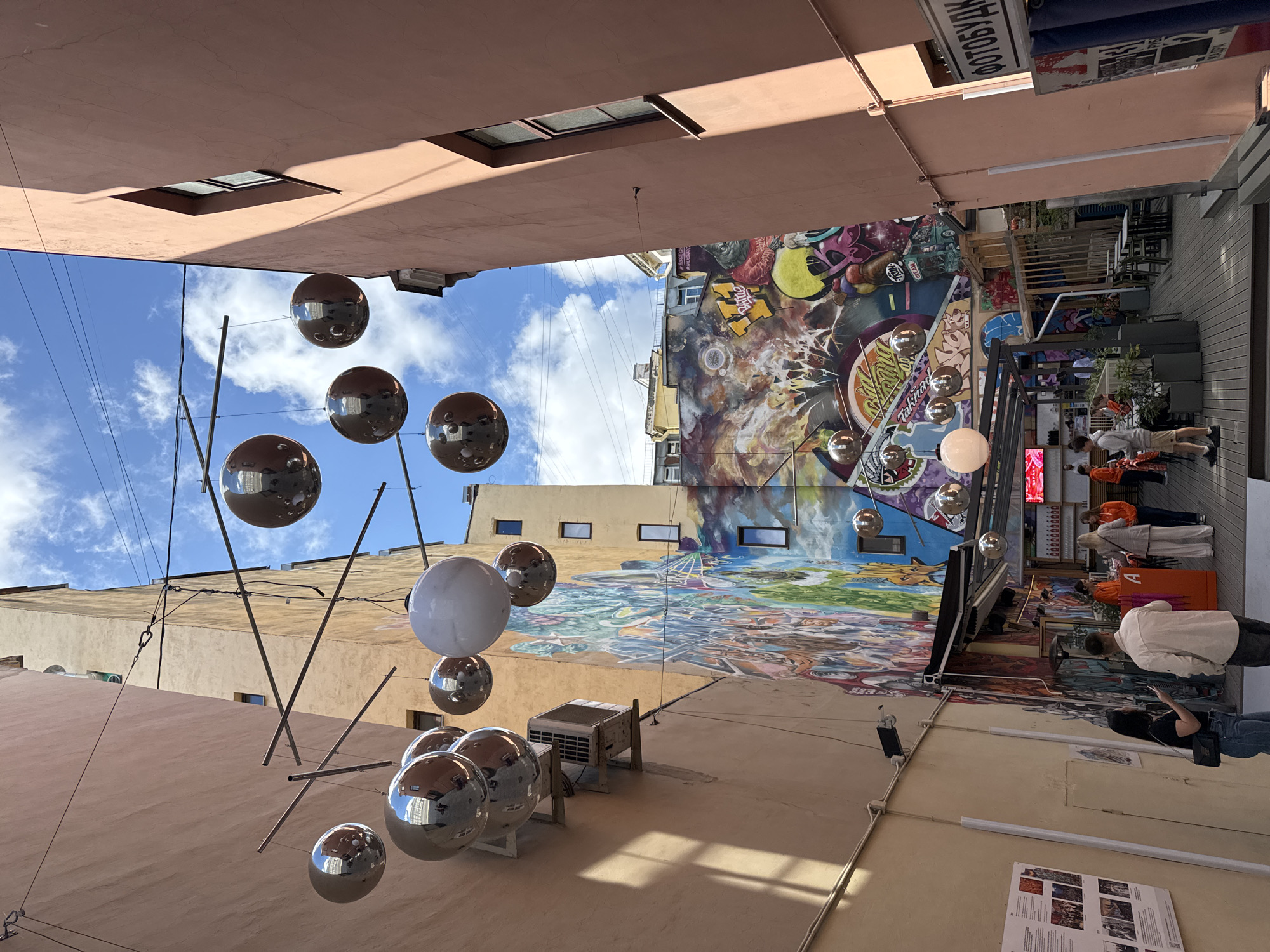 The image depicts a narrow street or alleyway in Saint Petersburg, Russia, lined with buildings featuring colorful graffiti murals. Suspended from wires above the alley are numerous reflective spherical decorations and one large white sphere. The alley opens up to a bright, partially cloudy blue sky.
On the left side, a peach-colored building wall is visible, with an air conditioning unit mounted on it, above which several of the reflective spheres are hanging. A large, intricate mural covers a significant portion of the wall further down the alley, depicting a vibrant and complex scene with figures, abstract shapes, and graffiti lettering.
On the right side, a light pink building with dark-framed windows is present. This building is partially in shadow. A sign with Cyrillic text is attached to its facade.
In the foreground and midground, several people are present, appearing to be patrons of an outdoor cafe or establishment set up within the alley. Tables and chairs are arranged on a wooden deck. Some individuals are standing, while others are seated. One woman in the foreground is wearing jeans and a black top, holding a phone. A man in a light-colored shirt and dark pants stands nearby. Other individuals are seen in the background, some wearing orange clothing.
The overall impression is that of a lively urban space, possibly a street art or cultural hub, with an artistic and festive atmosphere.