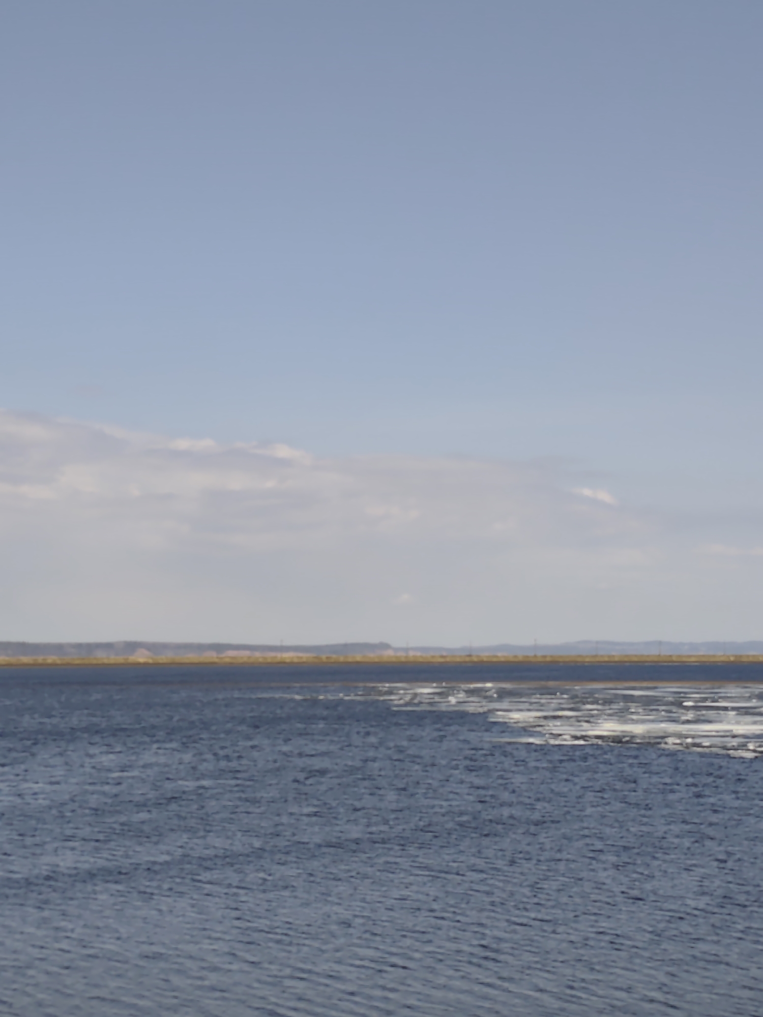 The image shows a body of water, likely the Kama River, with some ice floes near the shoreline. In the background, a distant landmass is visible under a cloudy, light blue sky. The location is likely near Chaikovsky, Russia, given the geographical features consistent with the Kama River and its surrounding landscape in that area.