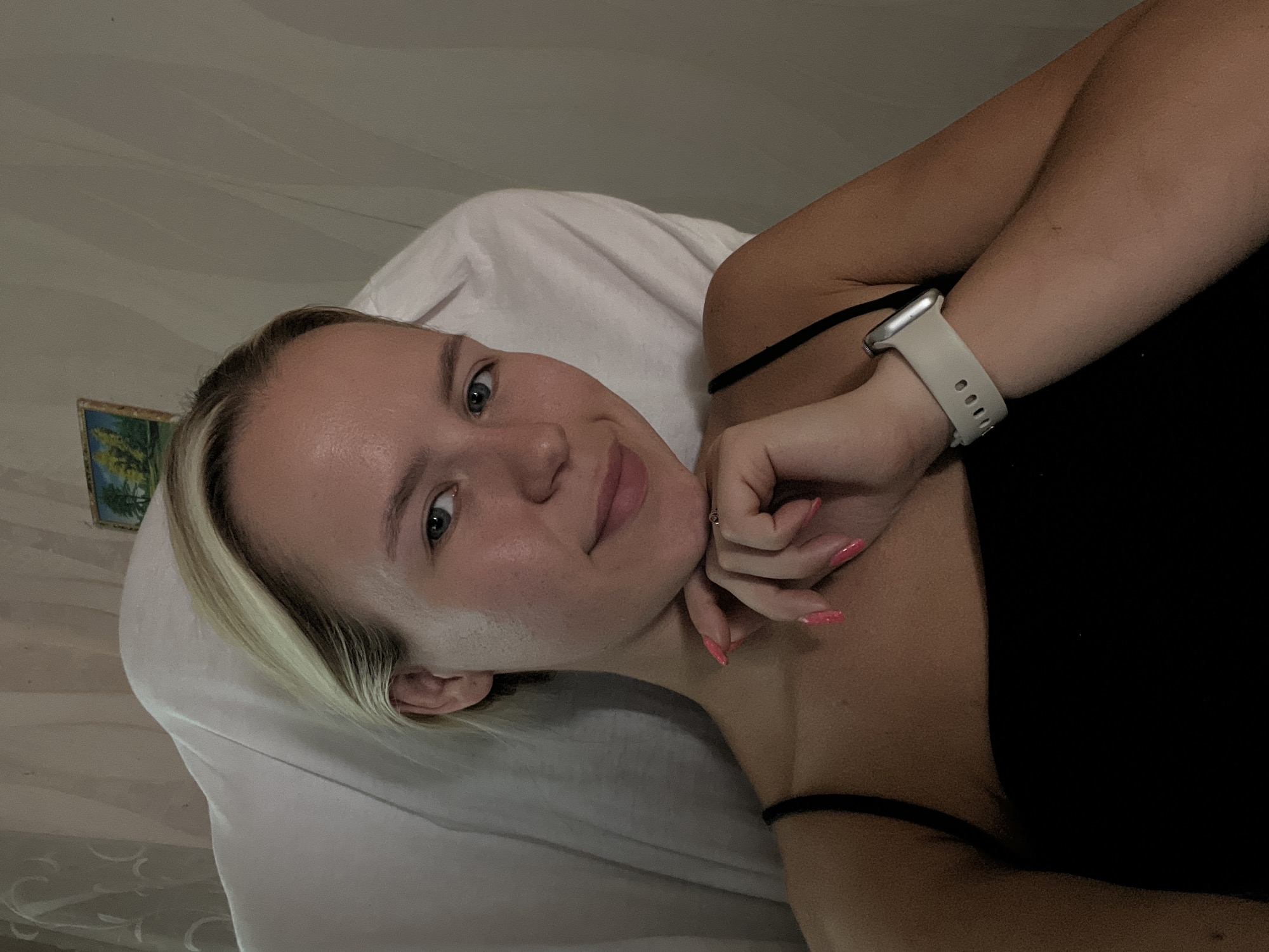 In the image, a young woman with blonde hair and blue eyes is positioned in the center, leaning back against a white pillow. She is wearing a black tank top and a silver smartwatch on her left wrist. Her right hand is resting on her chin, and her nails are painted a bright pink. A small framed picture hangs on the wall behind her. The lighting suggests an indoor setting, likely a bedroom or living room. The overall scene is casual and intimate.