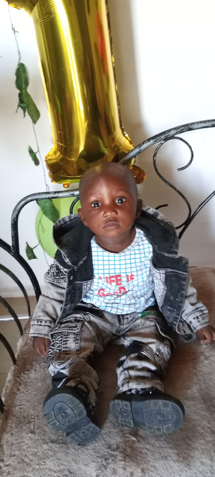 A baby sits on a chair. The child wears a blue plaid shirt with red lettering saying "LIFE IS GOOD", black and grey patterned pants, and a grey and black jacket. The baby has on dark-colored shoes. Behind the baby is a gold foil balloon in the shape of the number 1 and a green balloon. There is a climbing plant hanging down the wall. The location is likely a home in Jos, Nigeria, based on the background and subject ethnicity.