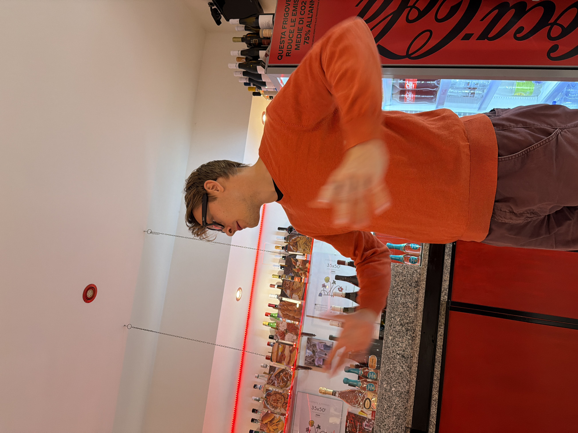 The image shows a man with glasses and an orange sweater standing in a shop, likely in Ragusa, Italy, based on the Italian writing on a cooler. He is facing a display shelf stocked with bottles of wine, and the cooler behind him has a Coca-Cola advertisement. The shelf seems to display advertisements and pictures of food, suggesting it might be a delicatessen or a specialty food store. The man's hands are slightly blurred, indicating movement. The lighting is bright, and the scene appears to be indoors.