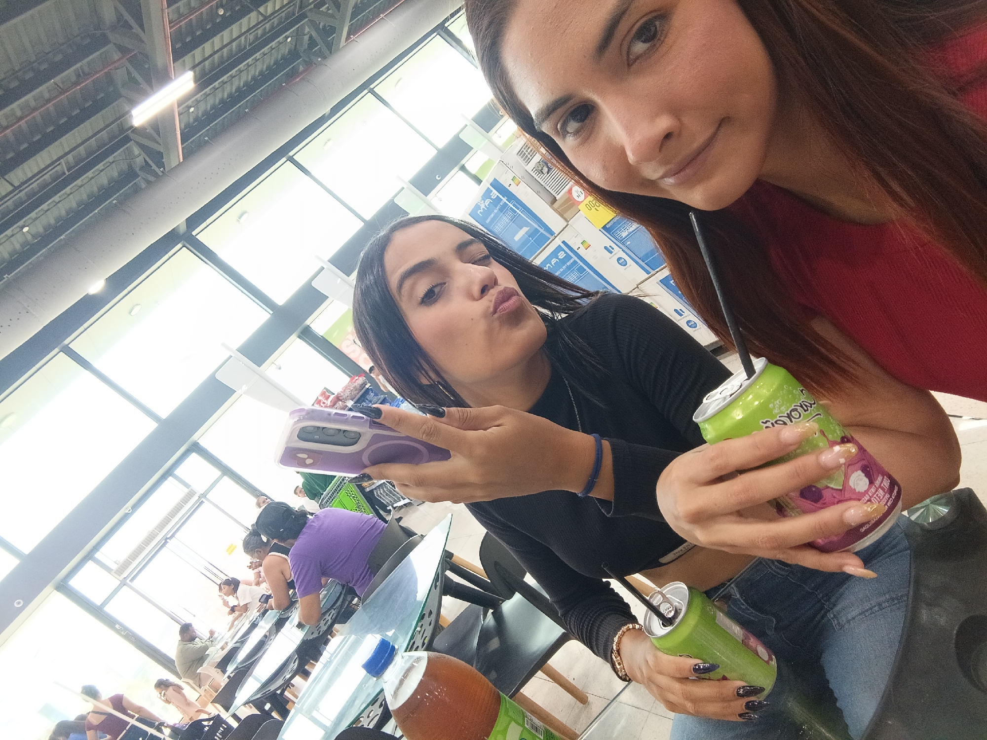 Two women are taking a selfie inside what appears to be a well-lit indoor space, potentially a cafeteria or eatery within a larger store or market. The woman on the left is holding a purple smartphone and making a puckered-lip expression. She has dark hair, a black long-sleeved shirt, and light-wash jeans. The woman on the right has auburn-brown hair and is wearing a red shirt. She is holding a green can with a straw. They are both sitting at a table. On the table, there is a glass bottle of juice, along with other chairs and people seated at tables in the background. In the background, there are boxes stacked. The location is Juan Griego, Venezuela.