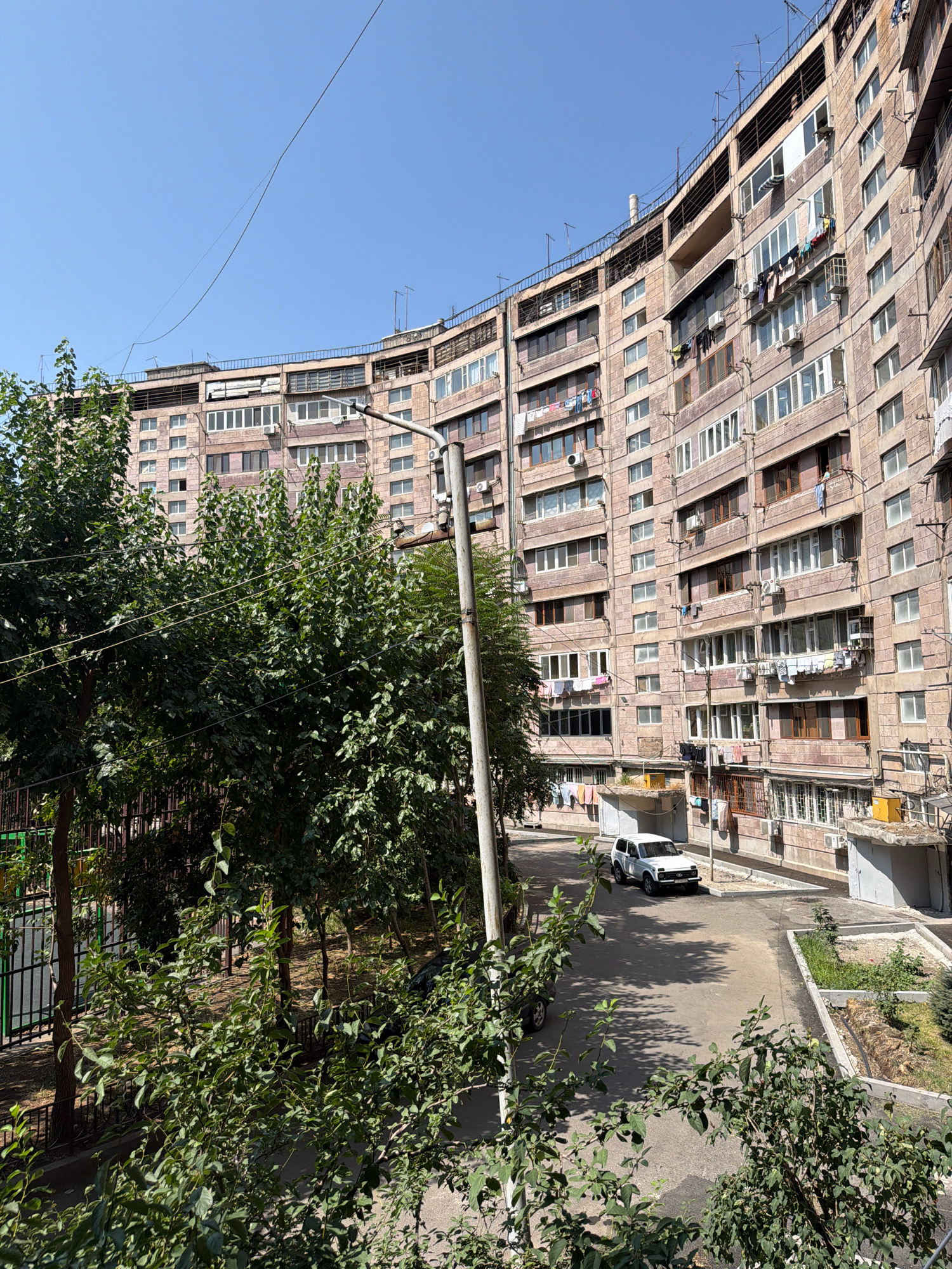 The image shows a curved, multi-story apartment building in Yerevan, Armenia. The building has a pinkish-brown facade and numerous balconies with laundry hanging out to dry. There are many windows, some with air conditioning units. A white SUV is parked on the asphalt road in front of the building. Trees and some greenery are visible in the foreground and to the left. The sky is clear and blue. The scene suggests a residential area in a city.