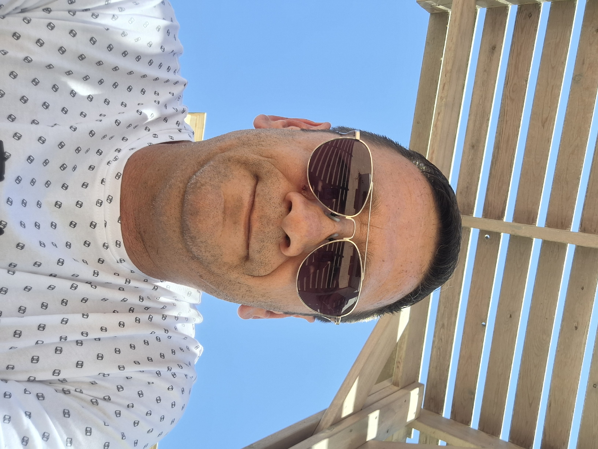 A man is seen from the chest up, wearing aviator sunglasses and a white t-shirt with a repeating pattern of small, dark-outlined cubes. He is positioned outdoors, under a wooden structure with horizontal slats, against a bright blue sky. The image is a selfie, as evidenced by the camera's perspective. The lighting suggests it is daytime. A portion of a building or structure with a light-colored facade is visible in the background, bottom left. The man's face is turned slightly towards the camera, and he has short, dark hair. The overall setting appears to be a relaxed, outdoor environment, possibly a patio or a shaded seating area. There are no other people or animals visible in the frame.