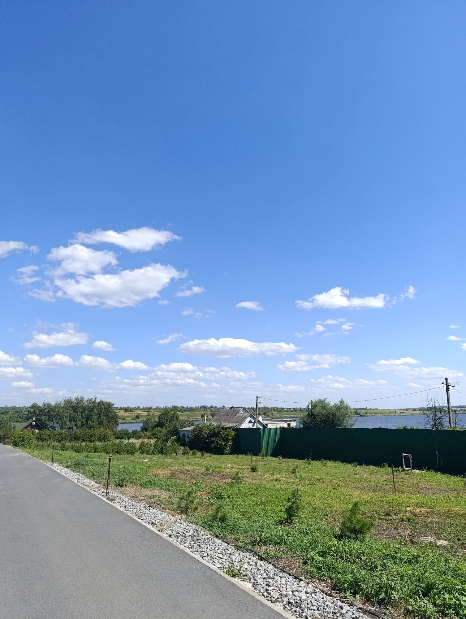 The image depicts a clear blue sky with scattered white clouds over a rural landscape. In the foreground, a paved road runs diagonally from the lower left corner into the scene. Alongside the road, a strip of gravel and then green grass. A dark green metal fence runs horizontally across the middle ground, separating a grassy field from a body of water in the background. Several trees are visible on either side of the fence, and a few small buildings, possibly houses, are situated near the water's edge. Power lines and poles are also visible in the distance. The scene suggests a sunny day in a tranquil, possibly residential, area.