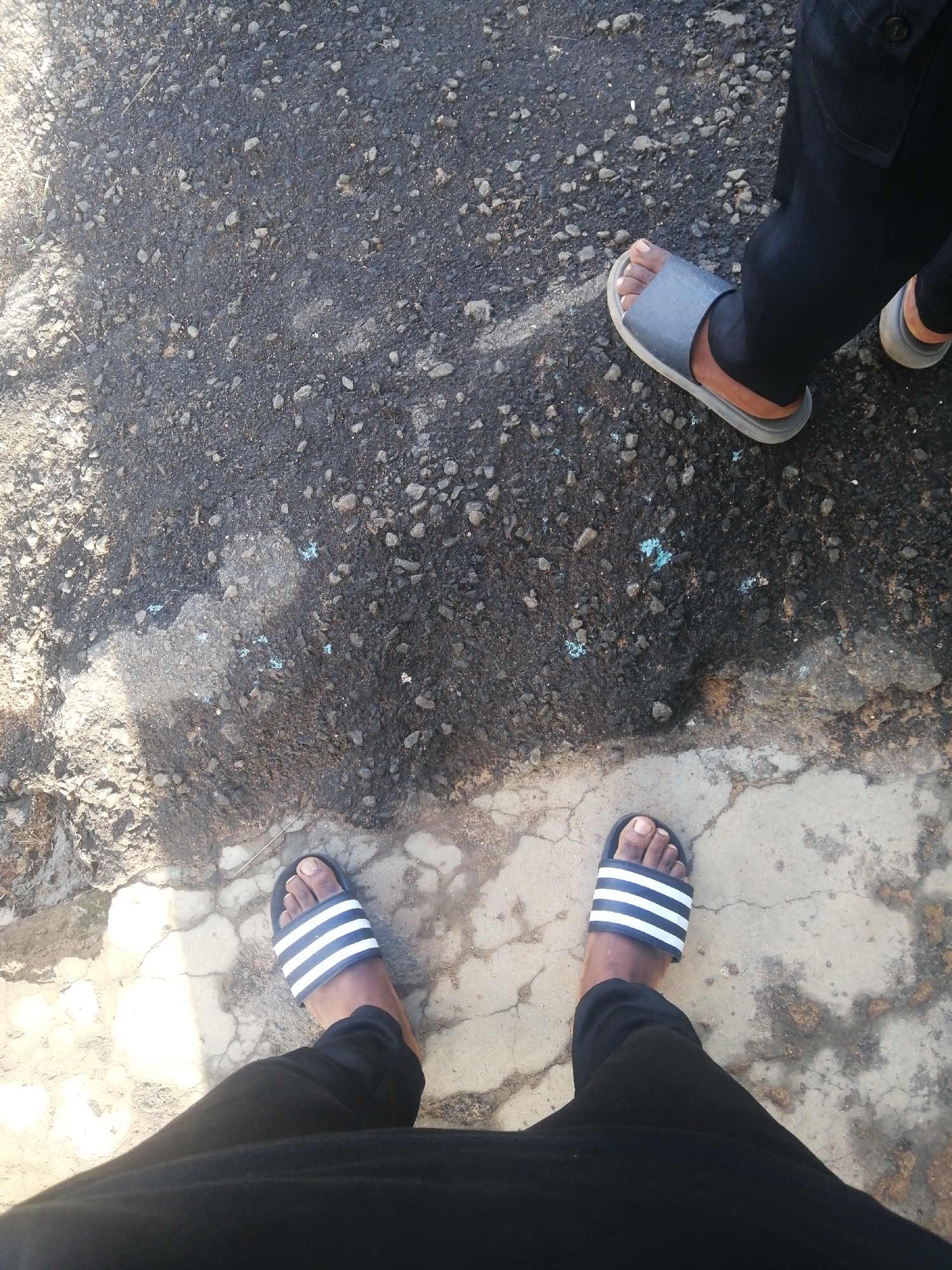 The image shows a person's feet and legs, wearing black trousers and sandals, standing on a rough asphalt surface with scattered gravel and some lighter concrete patches. There are two distinct sets of feet visible, suggesting two individuals are present, or one person has moved. One person is wearing grey slides, and the other is wearing black and white striped slides. The ground surface shows signs of wear and tear, with cracks visible in the concrete areas. The lighting suggests it is daytime, with shadows cast on the ground. The location is identified as Ajasse Ipo, Nigeria.