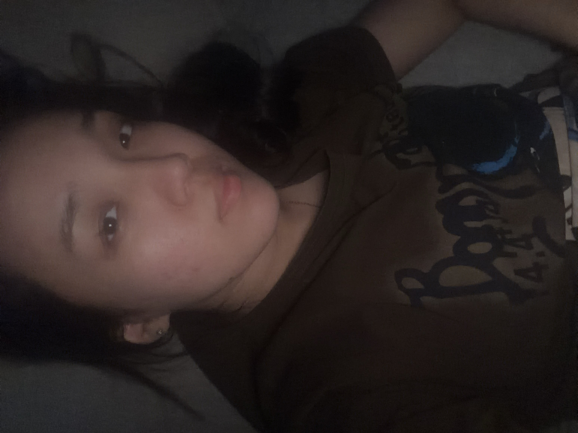 A young woman with dark hair and brown eyes is lying down, looking towards the camera. She is wearing a dark brown t-shirt with a graphic design on it. The lighting in the image is dim, with the majority of the scene in shadow, except for the woman's face and upper body which are illuminated. The background is dark and indistinct, suggesting an indoor environment. No specific location in the city of Unknown, Russia, can be determined from the visual information provided.
