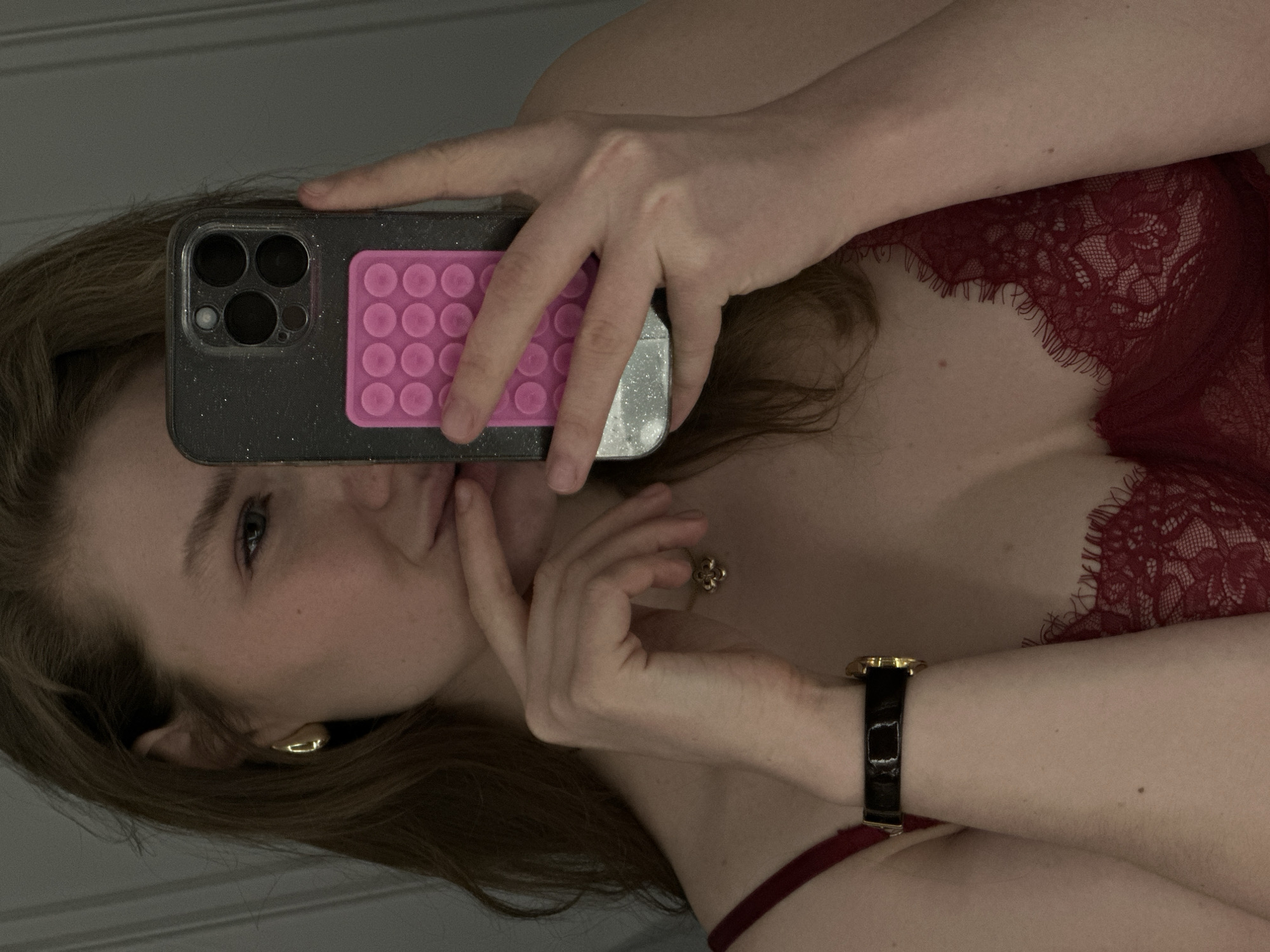A woman is taking a selfie in what appears to be a mirror. She is holding a smartphone with a pink pop socket on the back. The woman is wearing a red lace top and a black watch with a gold face. She has fair skin and brown hair. She is wearing gold earrings. The background is out of focus, but appears to be a neutral wall. There is no evidence to suggest this photo was taken in Kazan, Russia.