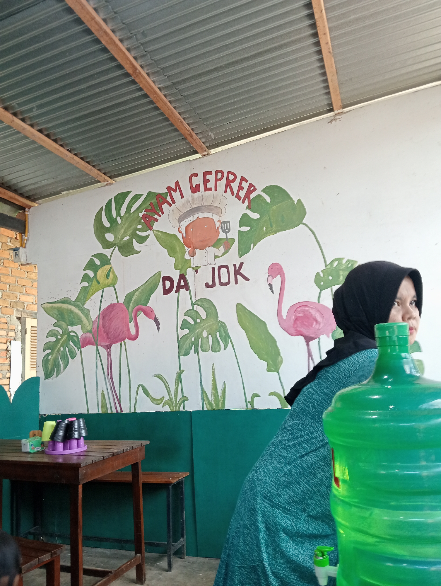 The image depicts an indoor scene, possibly a restaurant or eatery, with a wall mural. The mural features cartoonish depictions of tropical leaves, two pink flamingos, and a chef character. Text on the mural appears to read "AYAM GEPREK DA' JOK".
In the foreground, a person wearing a black hijab and a teal garment is partially visible, sitting next to a large green water dispenser. To the left, a wooden table and benches are set up, with a few cups and a pack of tissues on the table. The environment appears to be covered by a corrugated metal roof supported by wooden beams. Brickwork is visible on the left side of the image. The overall setting suggests a casual dining or waiting area in Campago, Indonesia.
