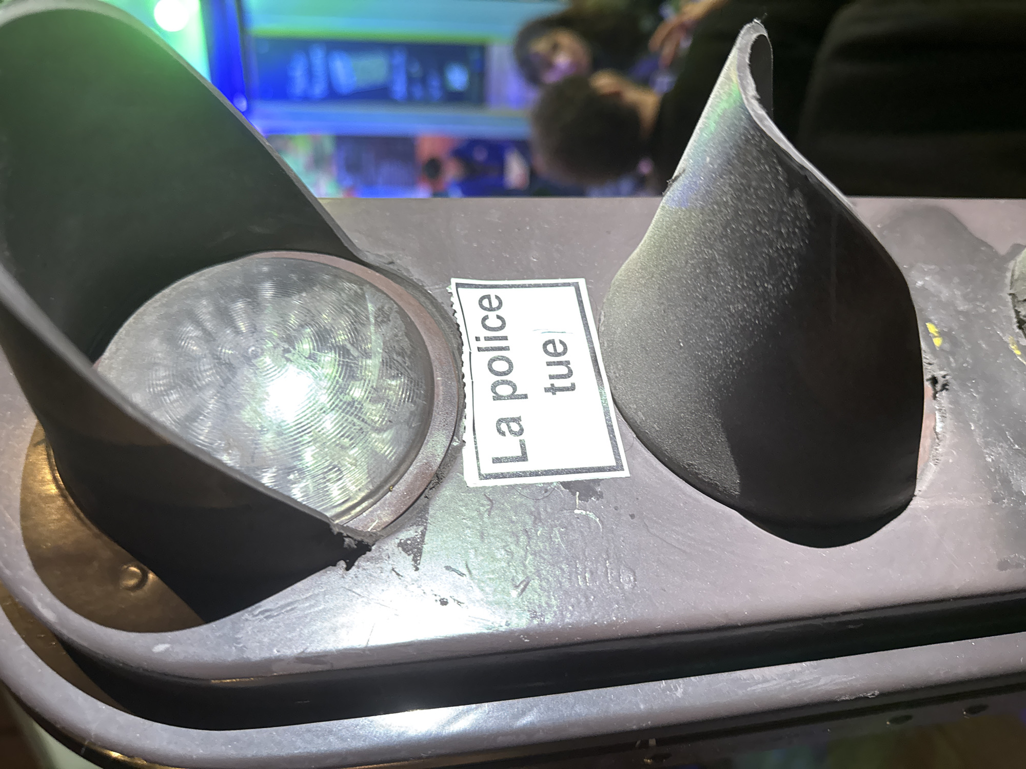 The image presents a close-up, high-angle view of a dark grey or black signal housing, likely part of a traffic light or pedestrian signal, in an urban environment. Two dark, conical hoods, designed to shield individual lights, protrude from the main unit. On the left, one circular light is partially visible, featuring a textured, possibly glass or plastic, surface with distinct concentric circles and a bright circular reflection from a light source.
Centrally positioned between the two light hoods is a rectangular white sticker with black text. The text reads vertically: "La police tue," which translates from French to English as "The police kill." The sticker appears to have slightly ragged edges, suggesting it was manually applied.
The background is heavily out of focus, showing blurred forms and colorful artificial lights in shades of blue, green, and purple. Indistinct outlines of people can be faintly discerned, suggesting a populated public space at night or in a brightly lit indoor setting. No specific activity is clearly visible beyond the placement of the sticker on the public infrastructure. The overall scene implies a form of public messaging or protest in an urban setting, consistent with the given location context of Paris, France.