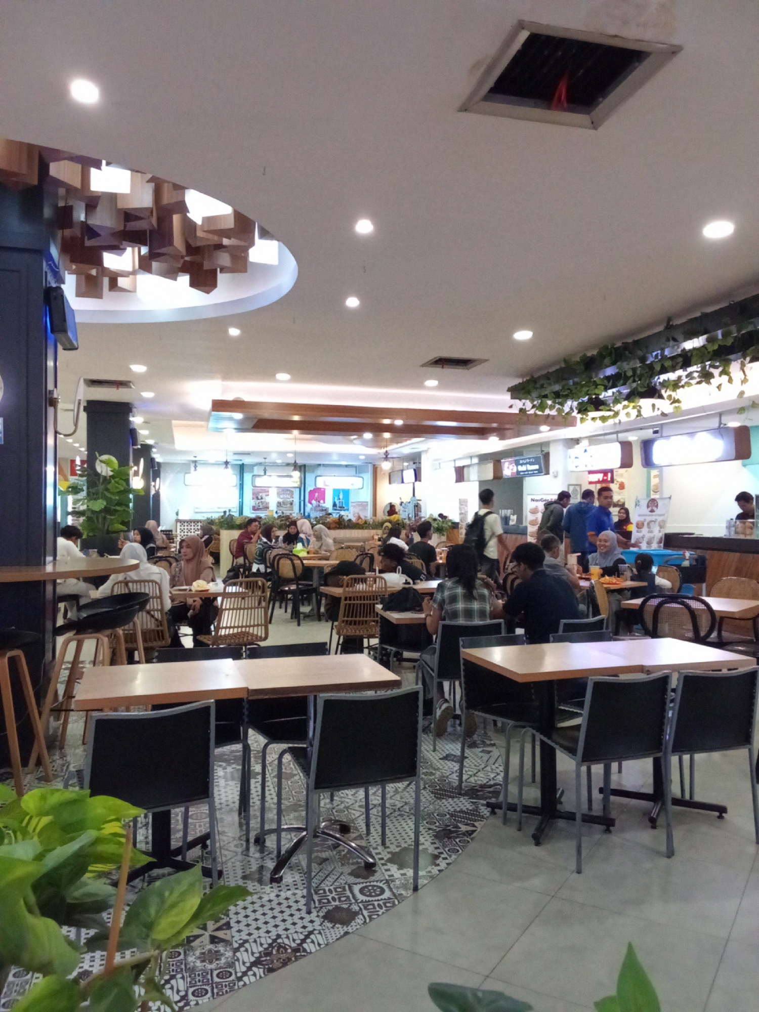 The image depicts a bustling indoor food court, likely located in Kediri City, Indonesia, given the context. The scene is populated with numerous individuals seated at various dining tables, engaged in eating, conversing, or waiting for their orders. The food court features a modern design with a high ceiling, incorporating ambient lighting and decorative elements such as wooden geometric installations and hanging artificial plants.
The seating arrangements consist of tables with dark-colored chairs, some of which appear to be office-style swivel chairs with wheels, while others are made of woven material. The flooring is a combination of tiled areas with intricate patterns and plain grey tiles. Various food stalls are visible in the background, some with prominent signage displaying menus and promotions, indicating a diverse culinary offering. People are dressed in casual attire, with many women wearing headscarves, suggesting a cultural context common in Indonesia. The overall atmosphere is one of a lively and functional dining space.