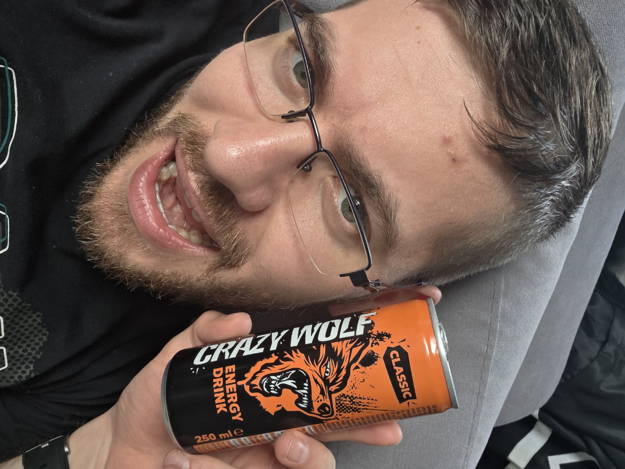 A man with a beard and glasses is holding a can of "Crazy Wolf" energy drink. He is lying down, possibly on a gray surface like a couch. The energy drink can is orange and black with a wolf graphic. The man is smiling with his teeth showing. The location of the scene in the city of Yagodovo, Bulgaria cannot be determined.