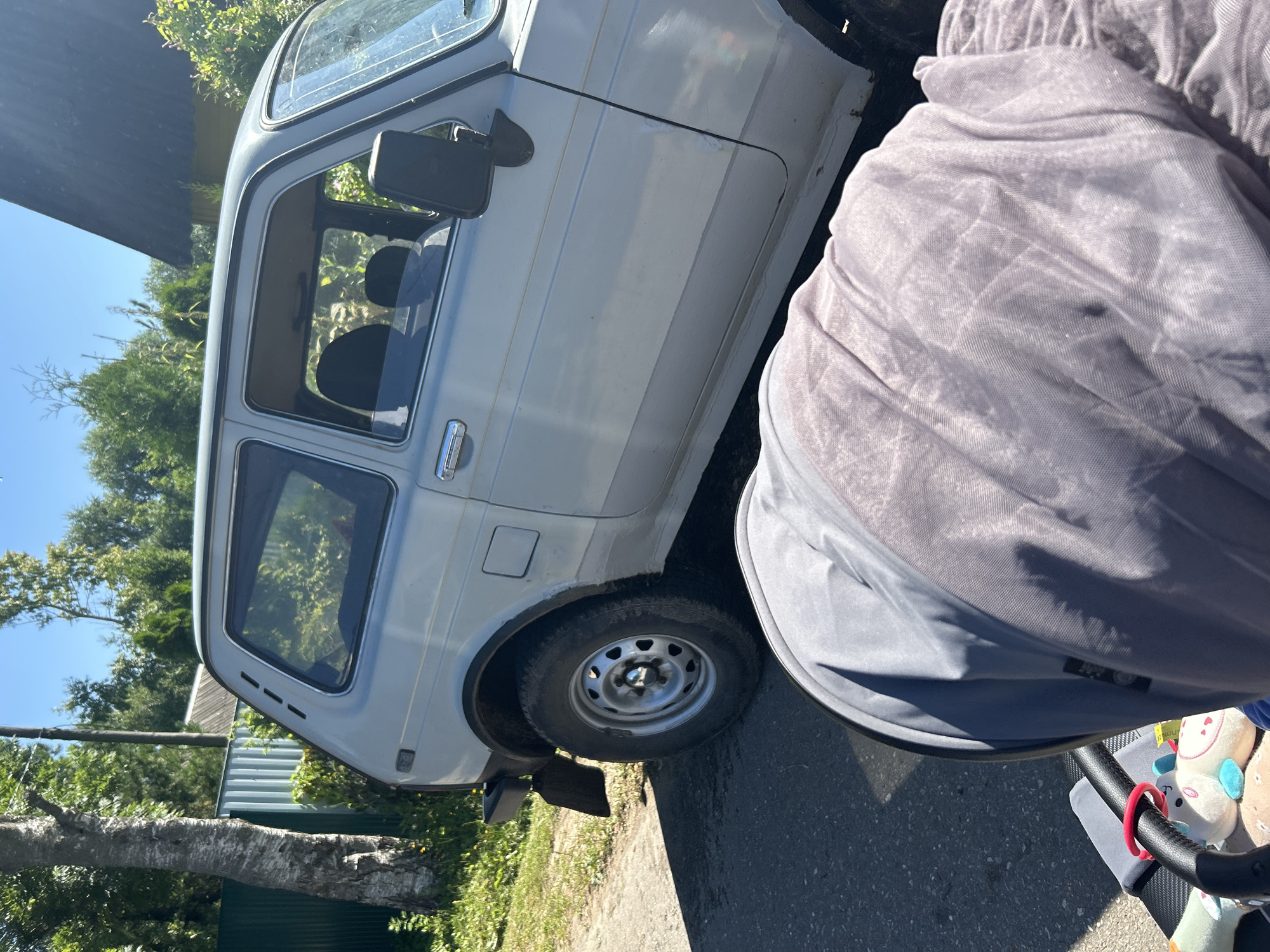 The image depicts an outdoor scene in the village of Kalitsino, Russia, on a sunny day. In the foreground, a baby stroller, covered with a light purple fabric, is positioned on an asphalt surface. The stroller has a black handle and several stuffed toys are visible inside. Behind the stroller, a light-colored, two-door car, possibly a Lada Niva, is parked on the side of the road. The car is light gray or silver and shows some signs of wear. The background includes a green corrugated metal fence, a tree with rough bark, and lush green foliage. A dark roof of a building is also visible in the upper right portion of the image, along with a utility pole. The lighting suggests it is daytime, with clear shadows cast by the car and the stroller.