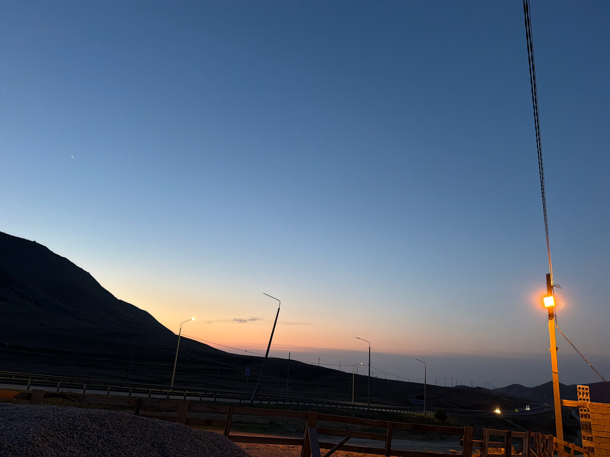 A wide-angle shot captures a desolate mountain landscape at dusk near Agachaul, Russia. The sky transitions from a deep blue overhead to soft hues of orange and pink near the horizon, where a setting sun is implied. A dark, silhouetted mountain range dominates the left side of the frame, its slopes textured by subtle contours. A highway runs horizontally across the middle ground, bordered by guardrails and illuminated by several streetlights that cast a warm glow. In the foreground, a pile of gravel sits behind a rustic wooden fence. A prominent lamppost with a bright orange light stands tall on the right, its wires extending upward into the twilight sky, where two faint celestial bodies are visible. The scene suggests a remote, undeveloped area transitioning into night, with the artificial lights of the highway providing the only significant illumination besides the dying light of day.