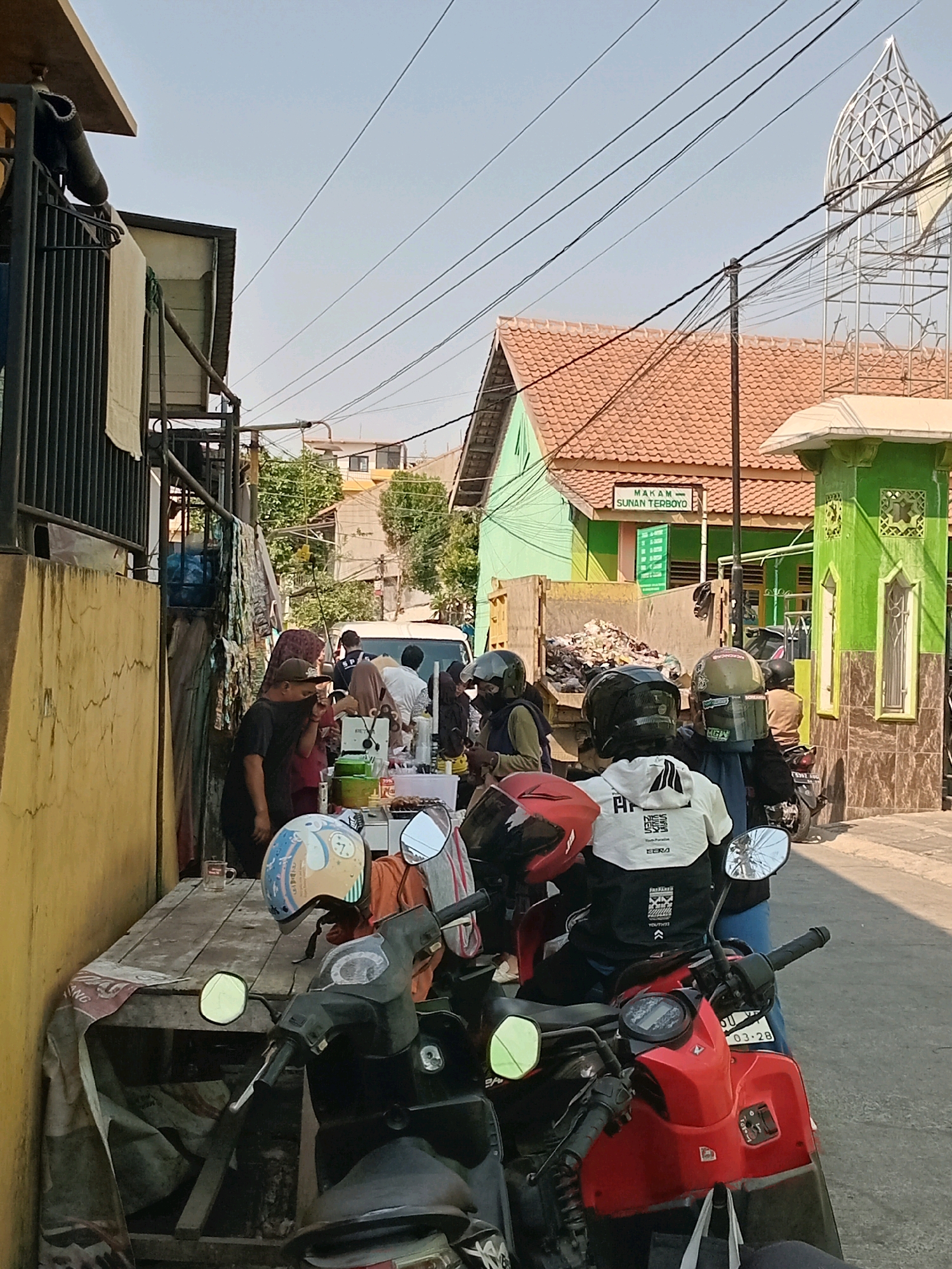 The image depicts a street scene in Semarang, Indonesia, likely in a residential or commercial area. In the foreground, several motorcycles are parked. A red motorcycle is prominent on the right, with a license plate reading "03-28". On the left, a dark-colored motorcycle is visible. People are gathered around a makeshift food stall or roadside vendor, which is set up on a wooden table covered with a patterned cloth. Several individuals, some wearing helmets, are seen interacting or queuing at the stall. They appear to be ordering or purchasing items. Behind the stall, a white van is parked. The surrounding buildings are typical of the region, with tiled roofs and brightly colored facades, including a green building with intricate architectural details and a sign that reads "MAKAM SUNAN TERBOYO". The sky is clear and blue, indicating fair weather. Overhead, power lines crisscross the frame, a common sight in Indonesian cities. The overall impression is a snapshot of daily life, with economic activity taking place on the street.