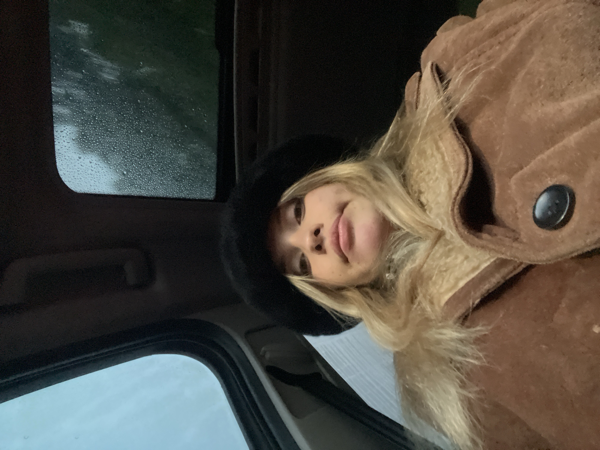 A woman with blonde hair and a black hat is seated inside a car. The car's window is visible, showing raindrops on the glass. She is wearing a brown suede jacket with a fuzzy collar and a button closure. The location is inside a vehicle, with the exterior potentially suggesting a rainy day, but there is no information on the specific location in the city of San Fernando del Valle de Catamarca, Argentina.
