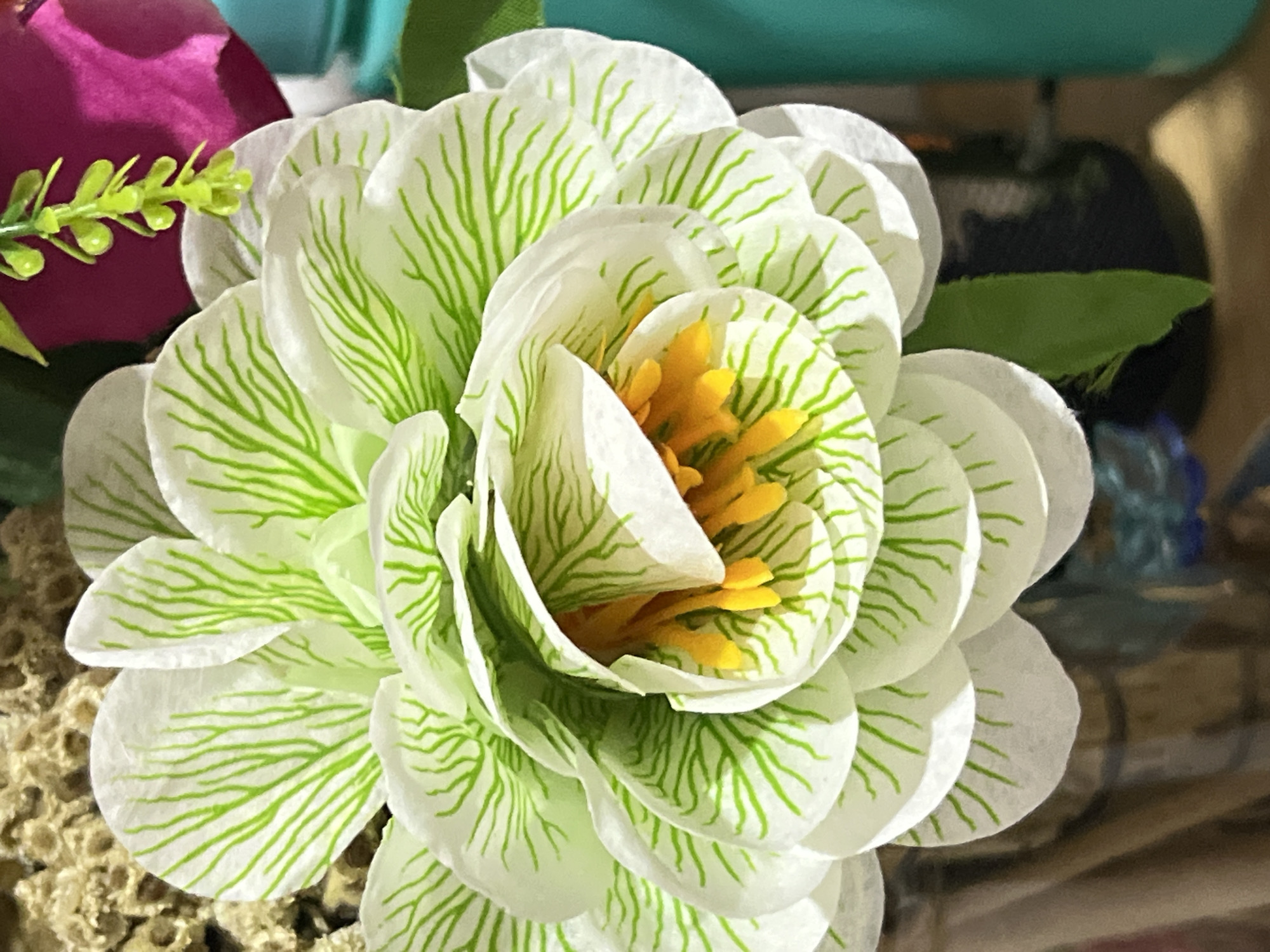 A close-up, eye-level shot focuses on a white artificial flower with green veining. The flower's petals are layered and appear to be made of fabric or plastic. The center of the flower reveals yellow stamens. In the background, parts of other artificial flowers, including a pink one and a green dangling sprig, are visible. The scene is set indoors, with the artificial flower arrangement likely placed on a table or shelf, possibly in a home or shop. The lighting suggests an indoor environment with artificial light or natural light filtered through a window. The image does not provide any information to indicate its location in Denpasar, Indonesia.